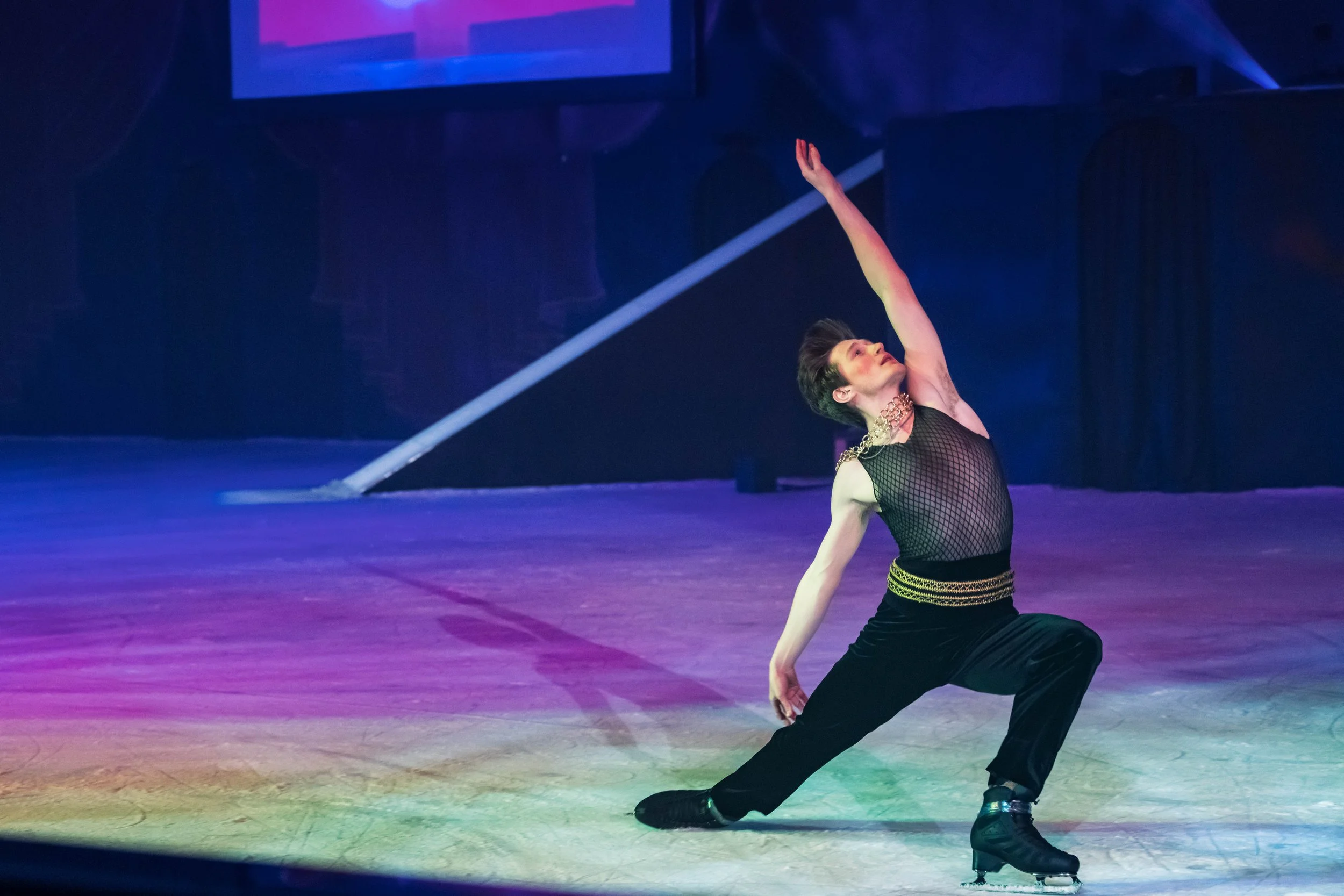 A male figure skater performing on ice, dressed in a black fishnet top with a choker and black pants, raising one arm and extending a leg in a dramatic pose.