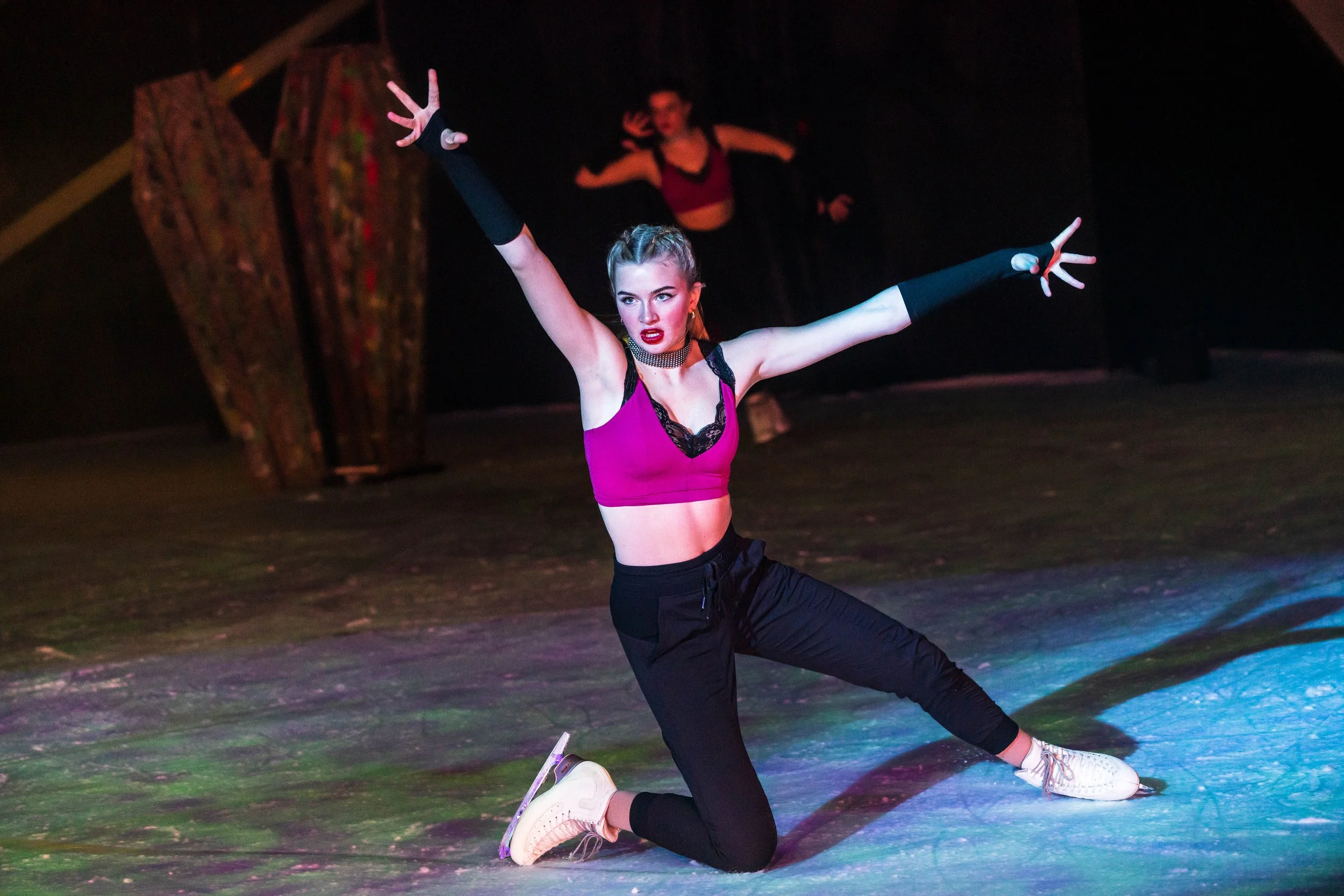 A woman in a purple sports top and black pants performing a dance move on stage with a dark background. She has her arms raised with hands open and appears to be in a dynamic pose, with a focused expression. A second woman is in the background with h