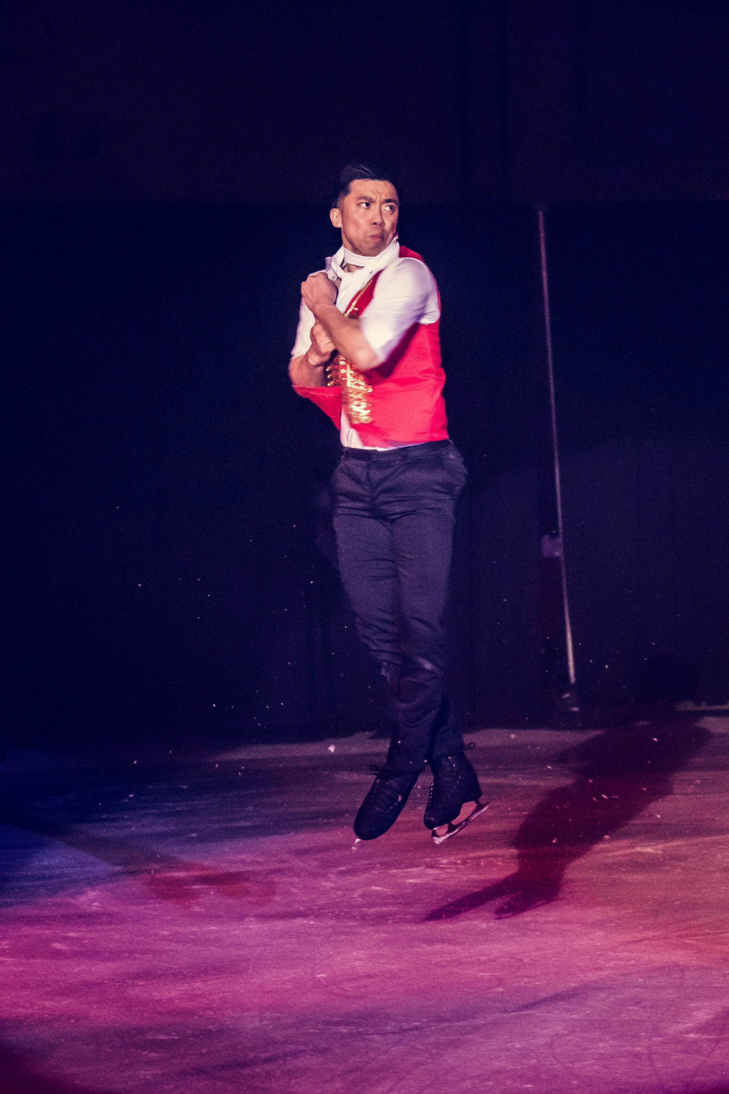 A man ice skating on a rink, wearing a red vest over a white shirt, with dark pants and bladed shoes. He has dark hair and appears to be in motion on the ice, with a dark background and red lighting.