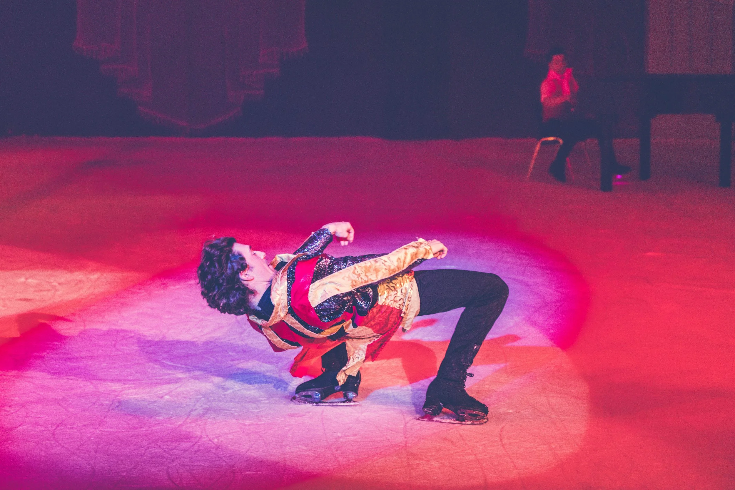 A performer lies on the ice rink in a dramatic pose during a show, dressed in colorful costume with black boots and ice skates, under pink and purple lighting with a person standing in the background.