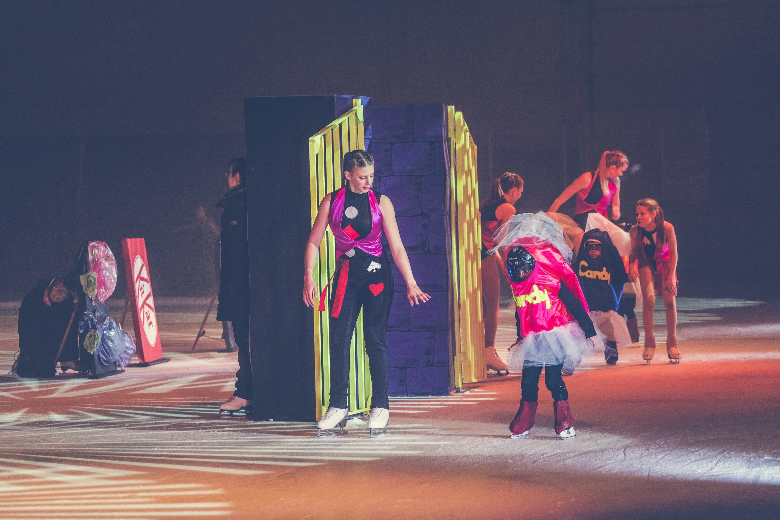 Group of young women in colorful costumes preparing on an indoor ice rink during a performance or rehearsal.