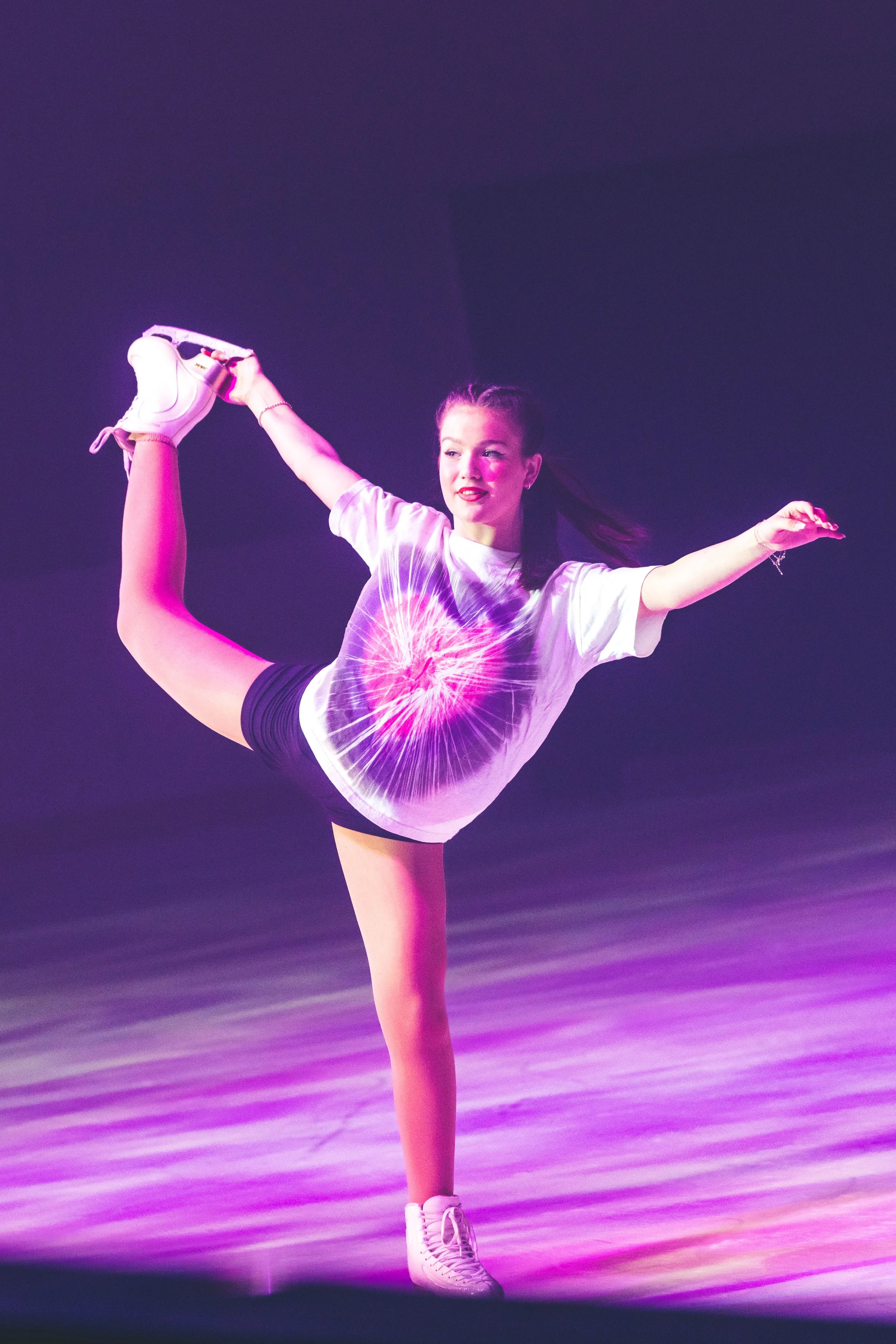 Performer in a white t-shirt and shorts executing an extended leg pose during a dance performance on a purple-lit stage.