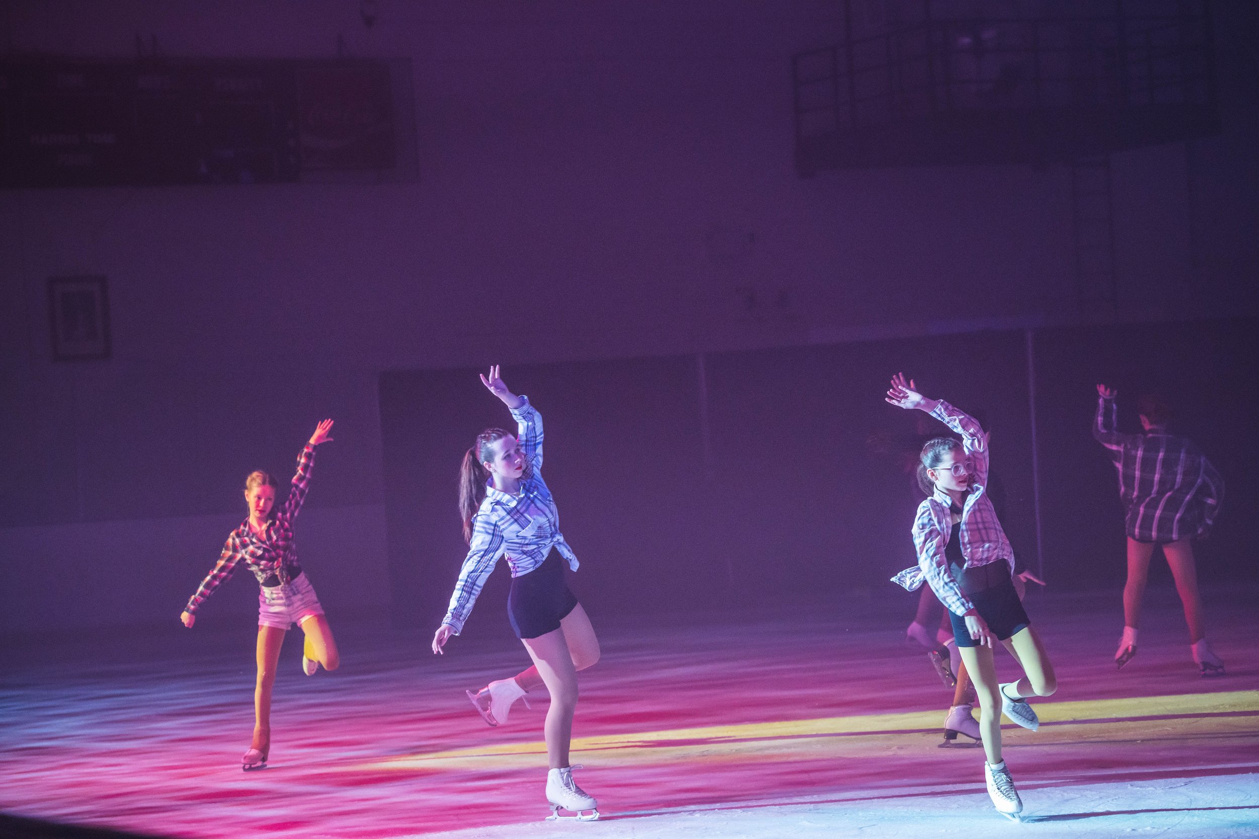 A group of young girls performing a synchronized dance routine on stage, wearing plaid shirts and skirts, with one girl raising her arm, under colorful stage lighting.