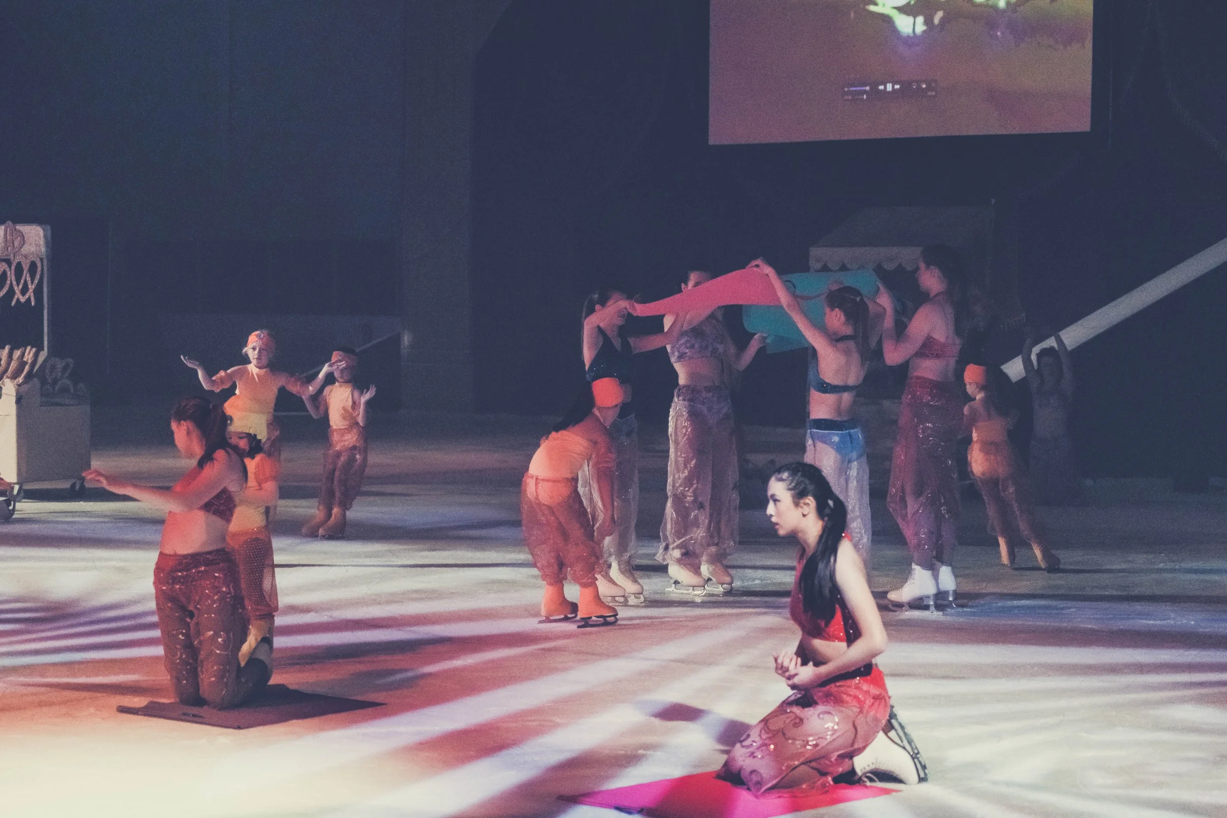 A group of young women practicing synchronized ice skating on an indoor rink, with some standing and others kneeling or sitting on the ice, wearing colorful costumes and skates.
