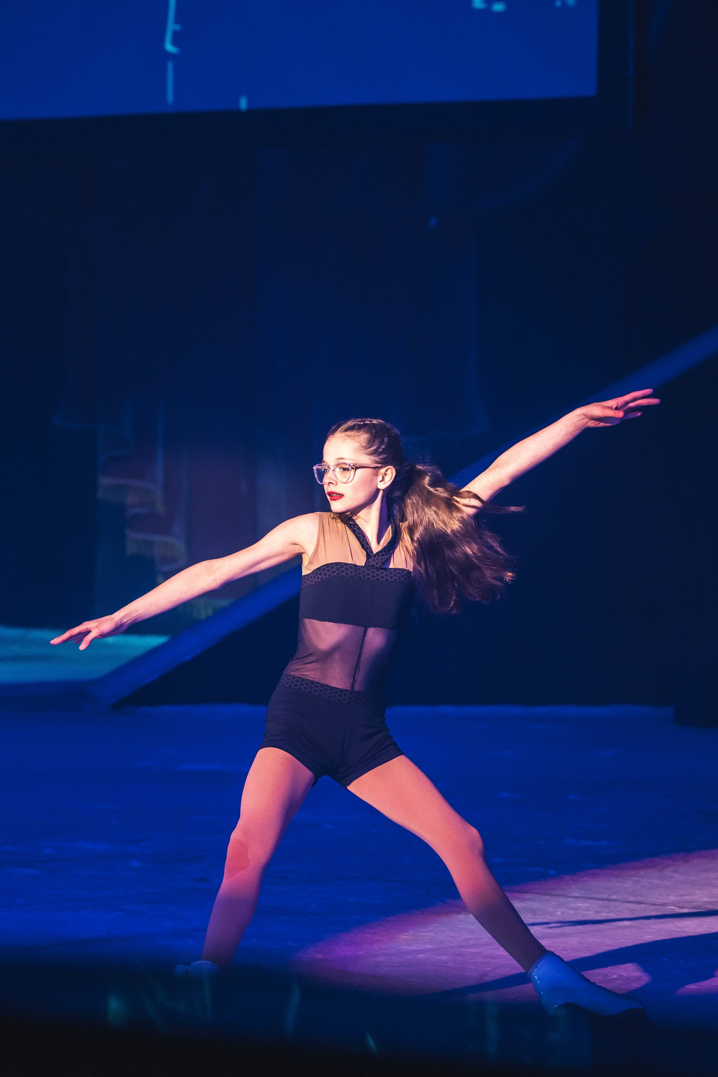 Young female ballet dancer performing on stage with arms extended, wearing glasses, black and brown dance costume, with stage lighting.