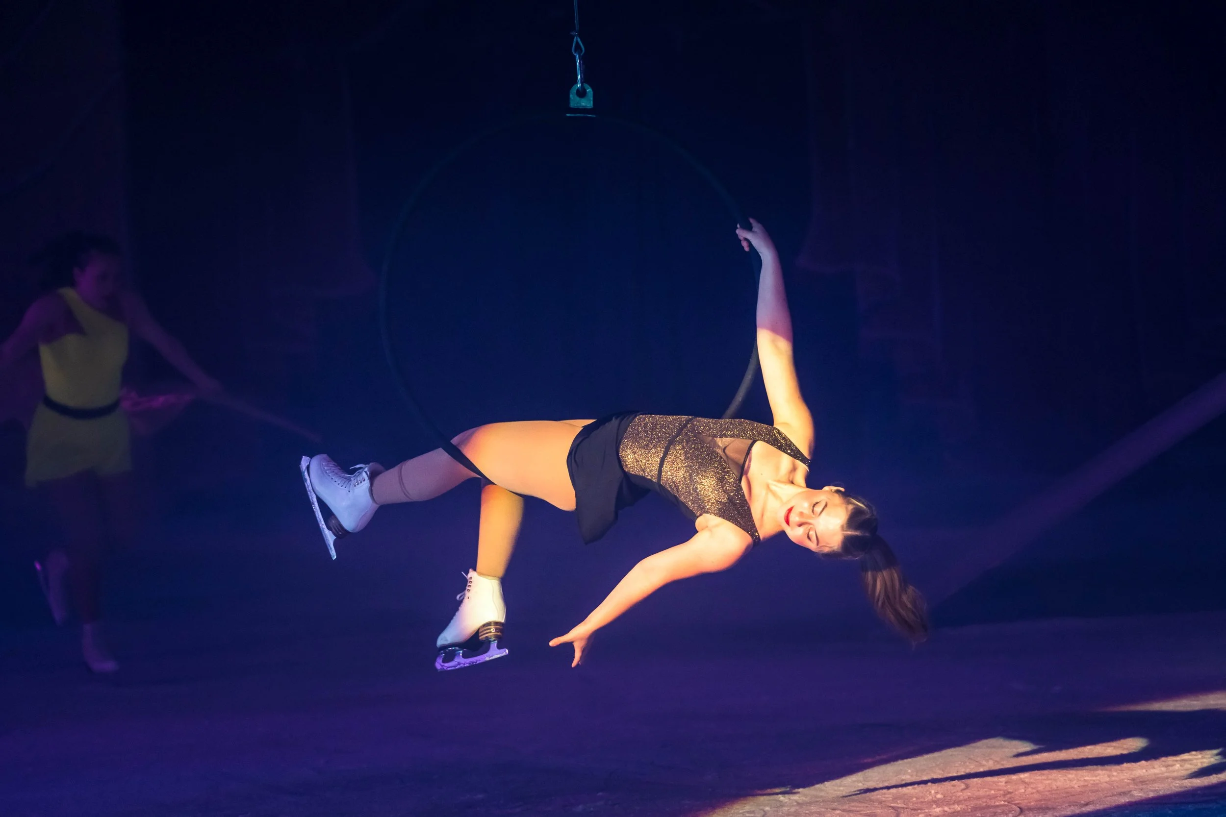 A female ice skater in a black and gold outfit performing on ice with a partner supporting her, surrounded by purple stage lighting.