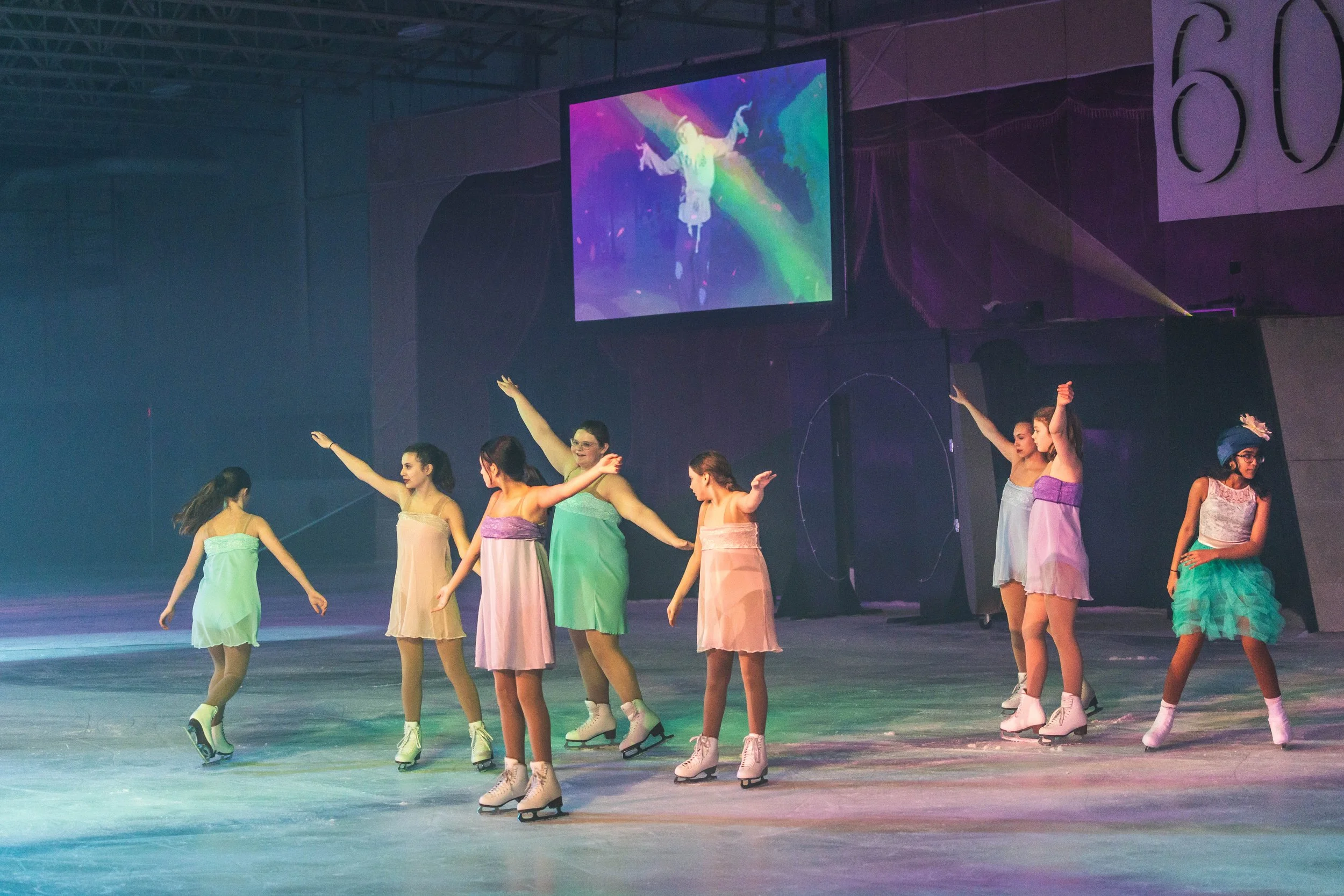 A group of young female figure skaters wearing pastel-colored dresses skating together on an ice rink during a performance, with a large screen displaying a colorful graphic in the background.