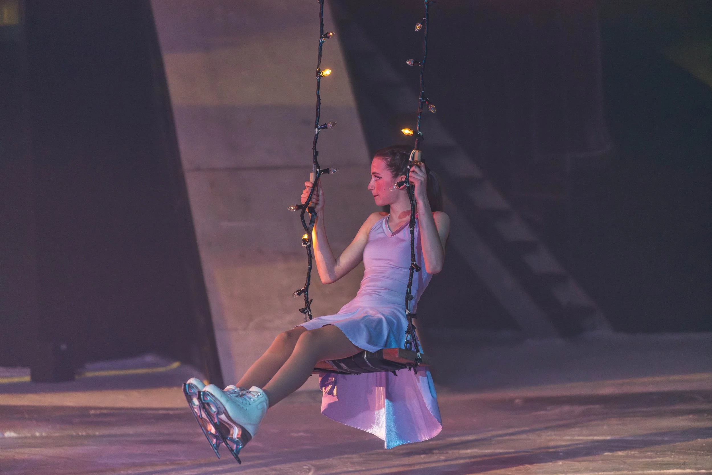 A young woman wearing a white dress and rollerblades sitting on a swing made of string lights with black guidelines, in an indoor setting with dim lighting.