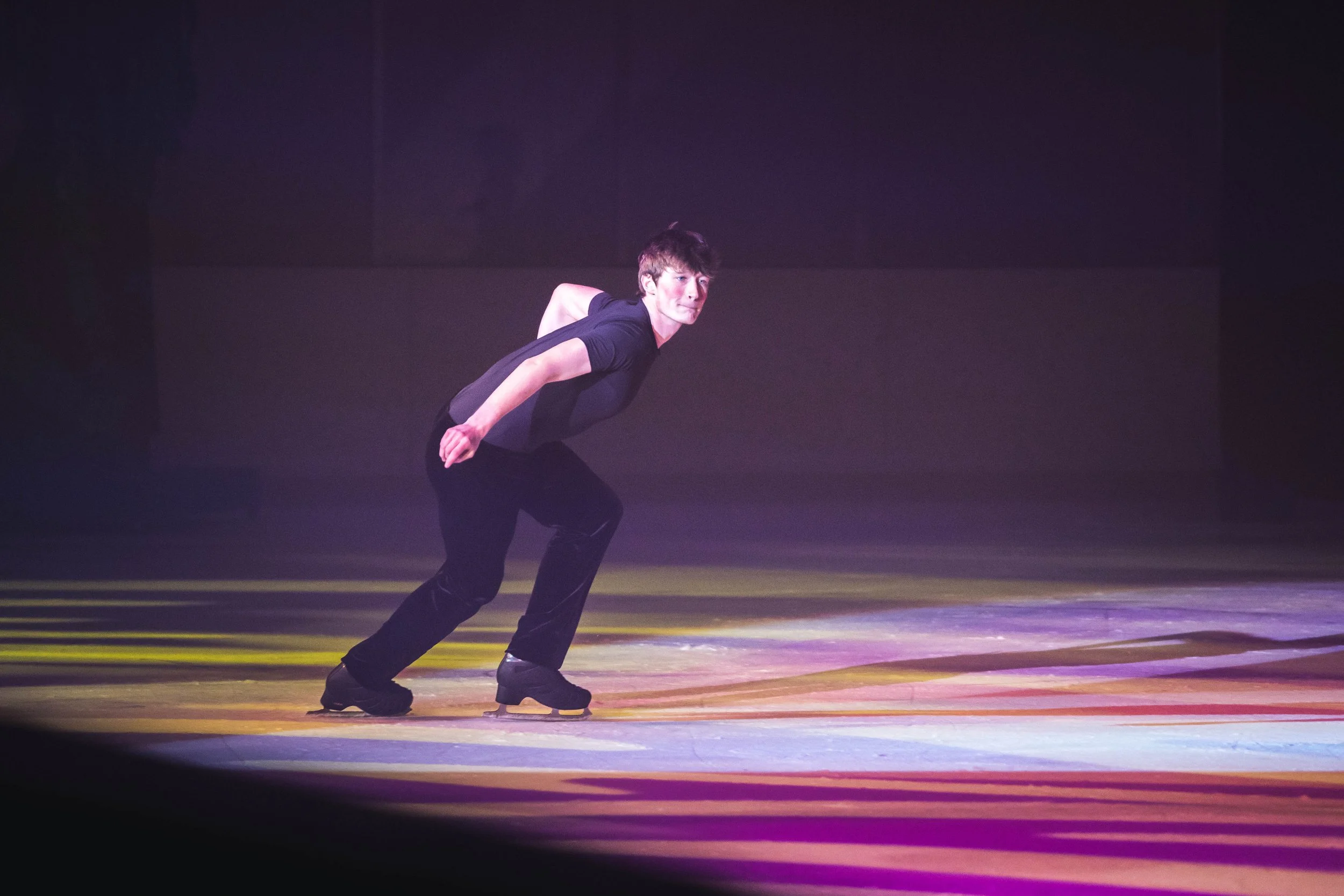 A person in a black shirt and black pants skating on a wooden floor, with a dark background, illuminated by stage lights.