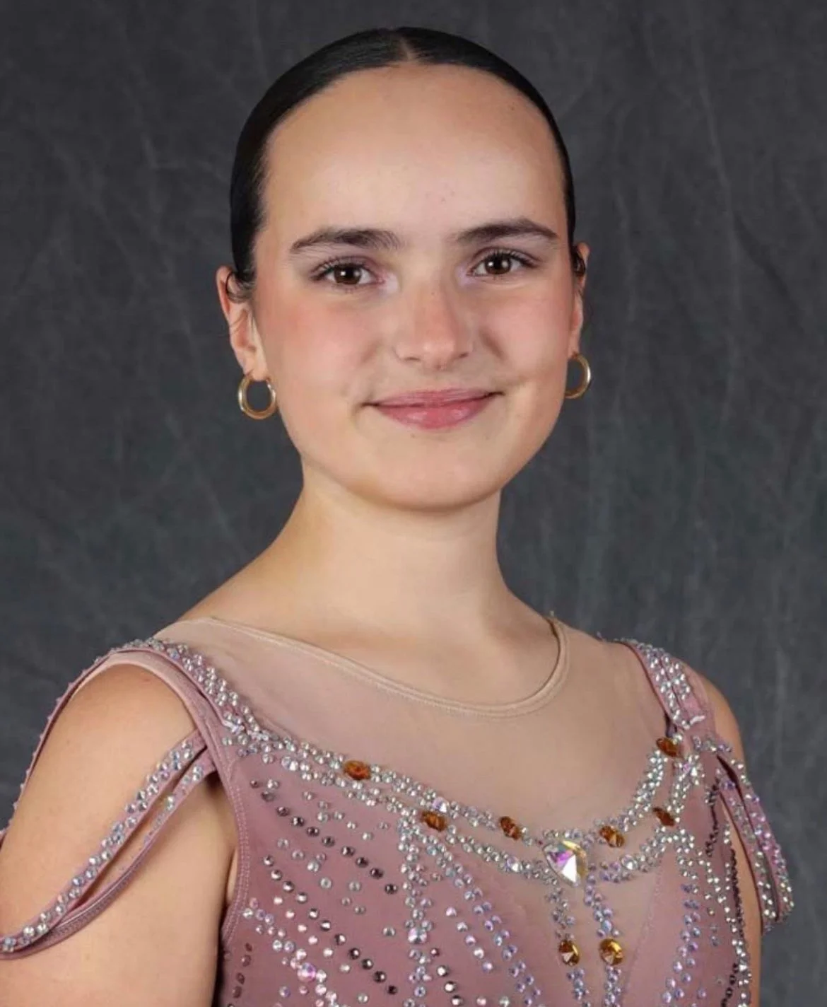 Portrait of a young woman with dark hair, wearing a pink dress embellished with rhinestones and earrings, smiling in front of a dark gray background.