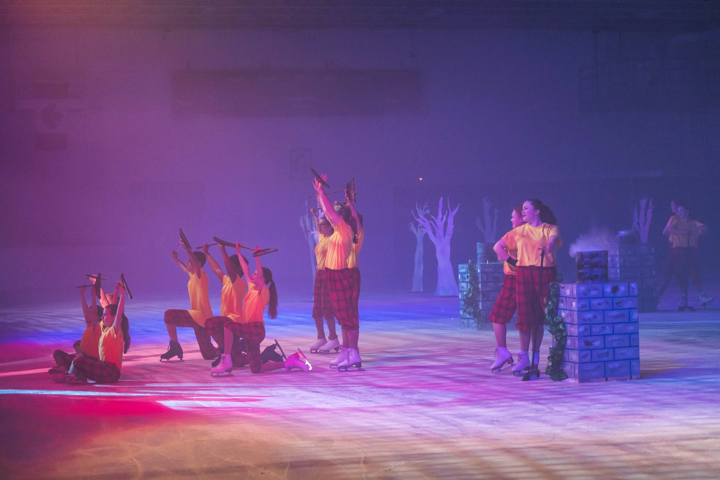 A group of performers on an ice rink, dressed in yellow shirts and red plaid pants, some standing and others kneeling, holding skate brushes in the air. The background features white trees and blue lighting, creating a theatrical atmosphere.