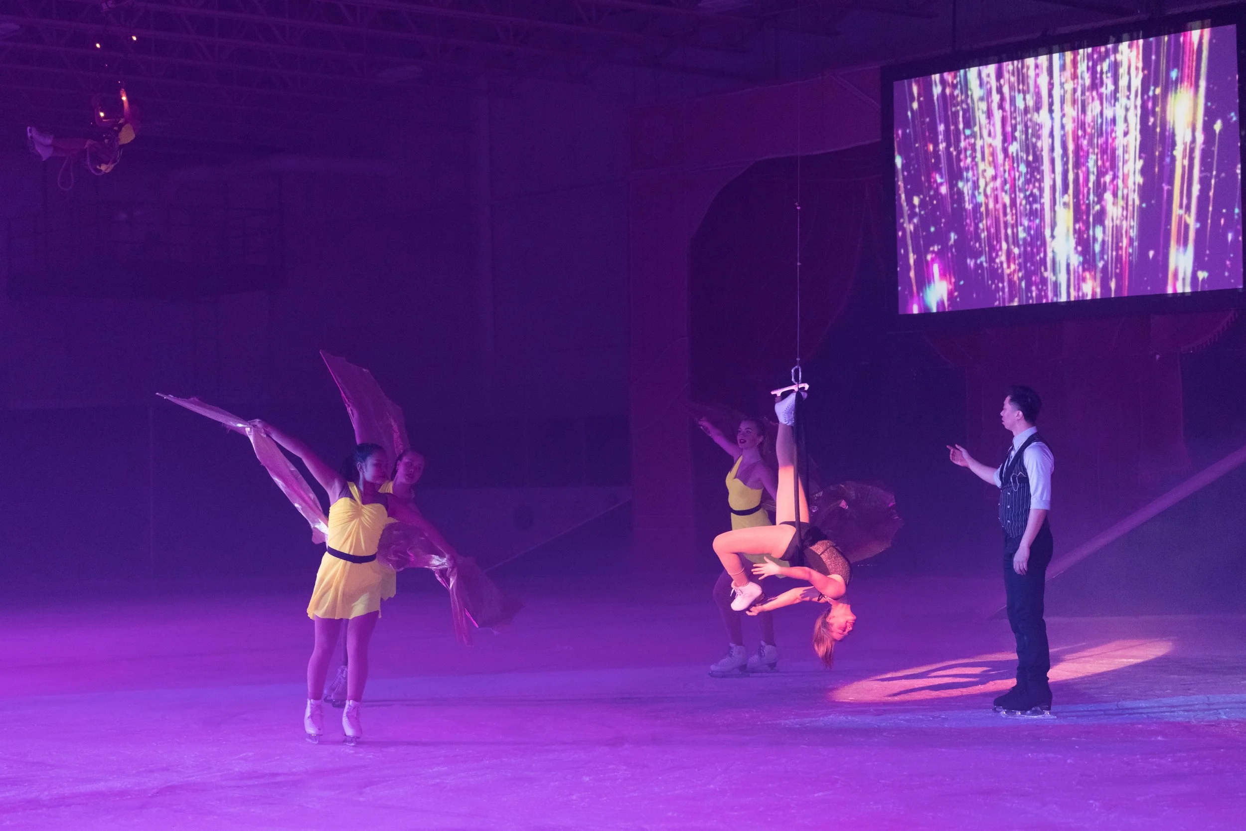 Performers on stage in yellow costumes with purple lighting, some acrobatic acts, and a large screen displaying colorful visual effects in the background.