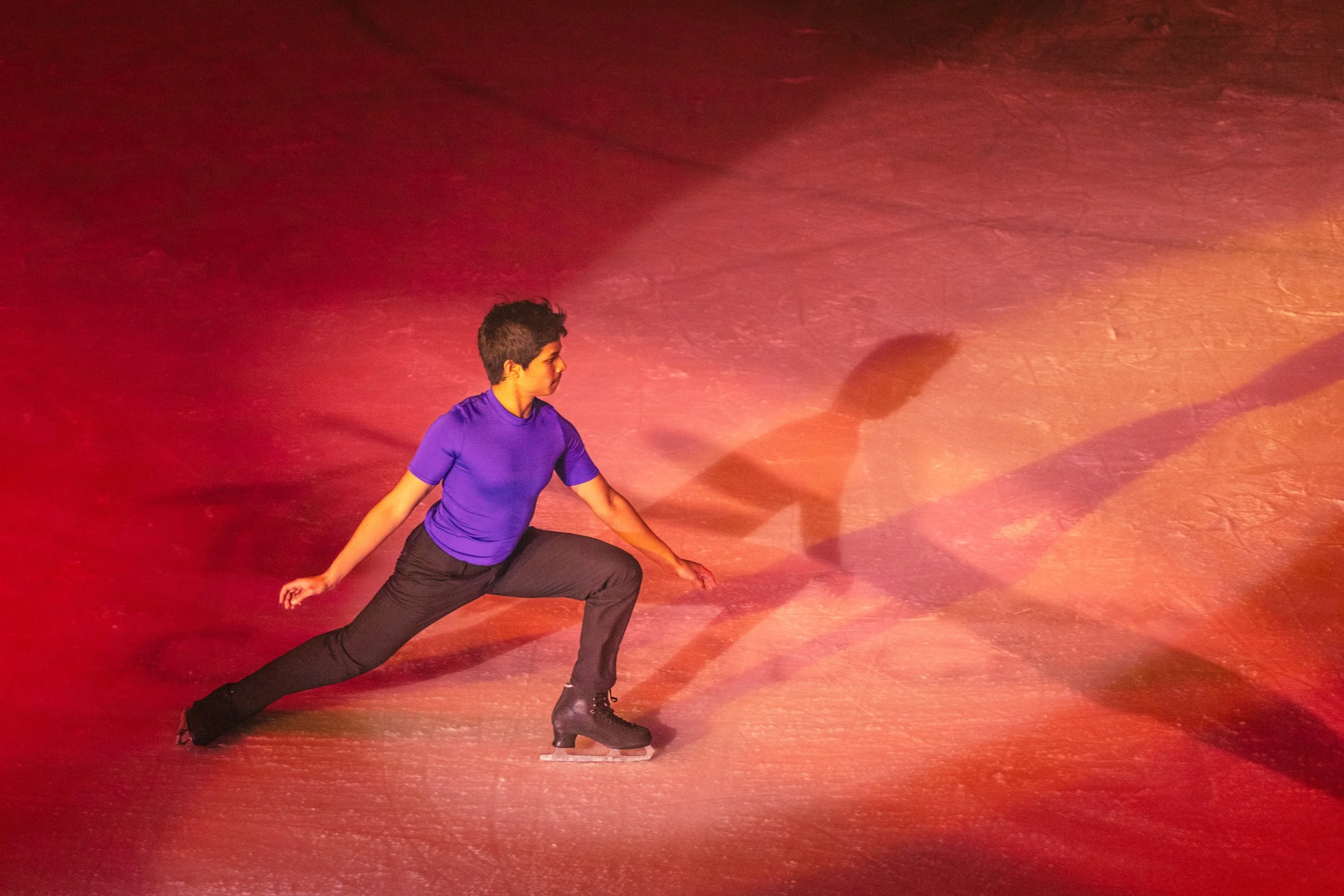 A person practicing figure skating on an ice rink in a deep lunge pose, wearing a purple shirt, black pants, and ice skates, with warm lighting casting shadows on the ice.