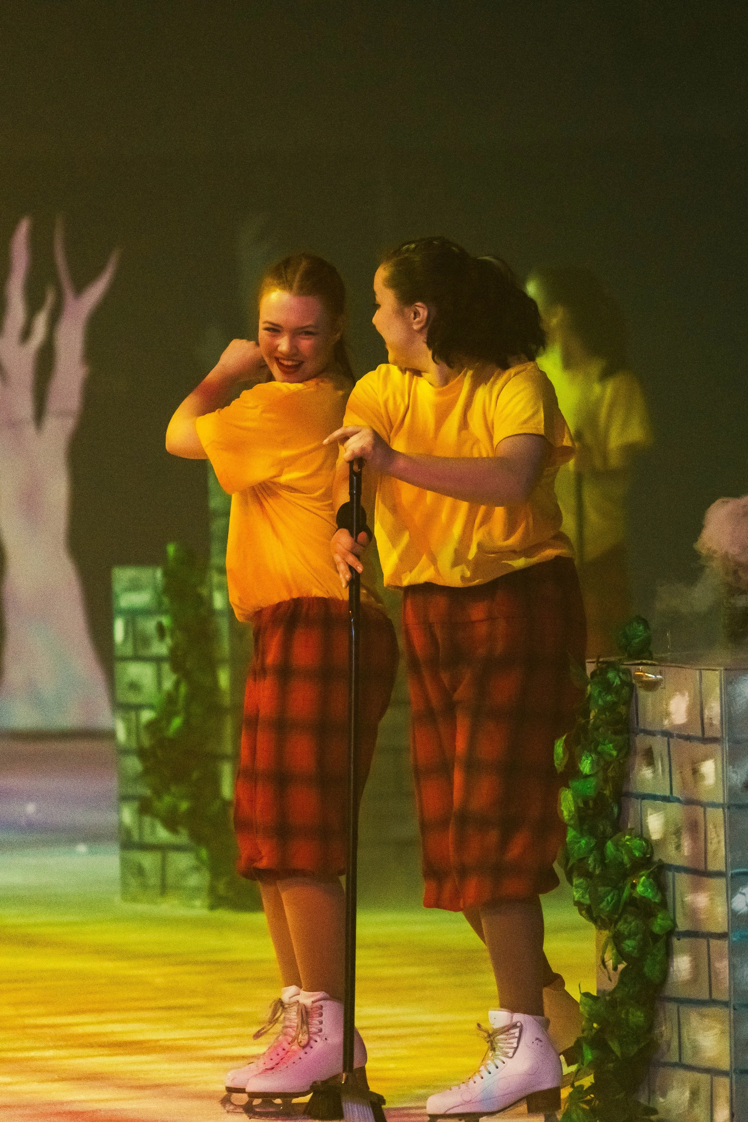 Two women on ice skates wearing yellow shirts and red plaid skirts, standing near a microphone on a stage with a green and yellow background, smiling and laughing.