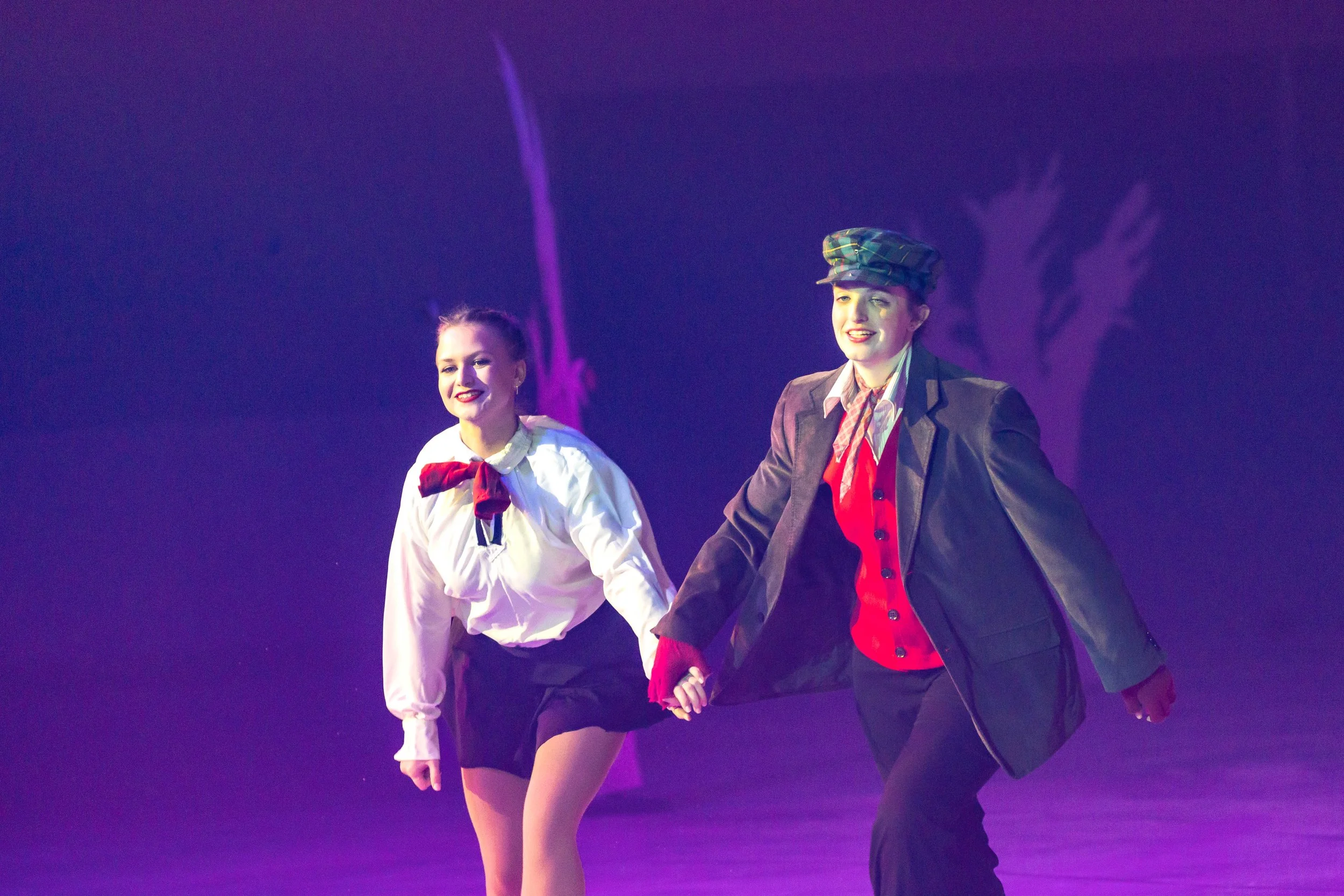 Two young women perform a dance routine on stage. One wears a white top with a red scarf, and the other wears a blazer, plaid hat, and red vest. They hold hands and smile while dancing under purple stage lighting.