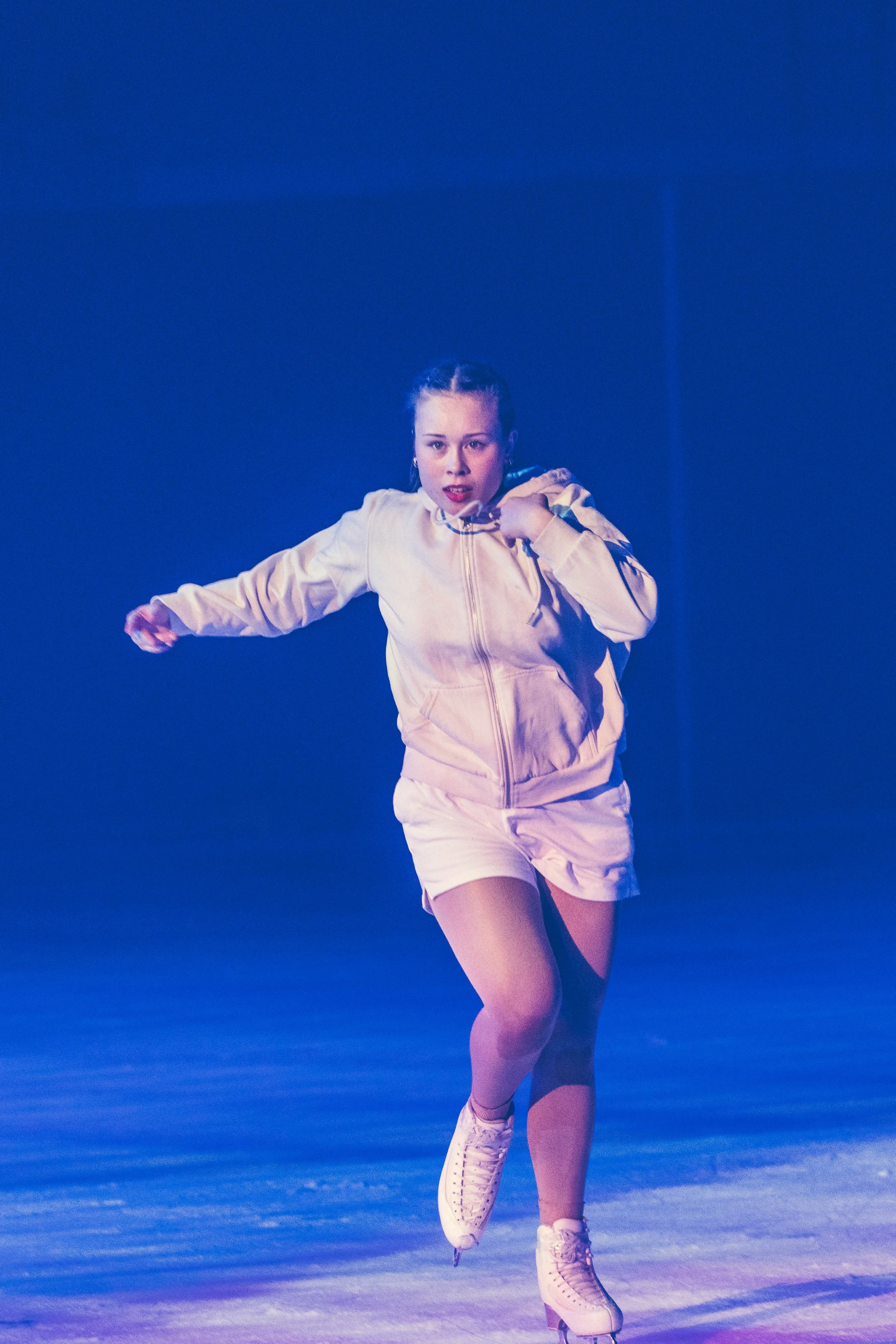 Young woman ice skating on a rink with a blue background, wearing a light pink jacket, matching shorts, and white ice skates.