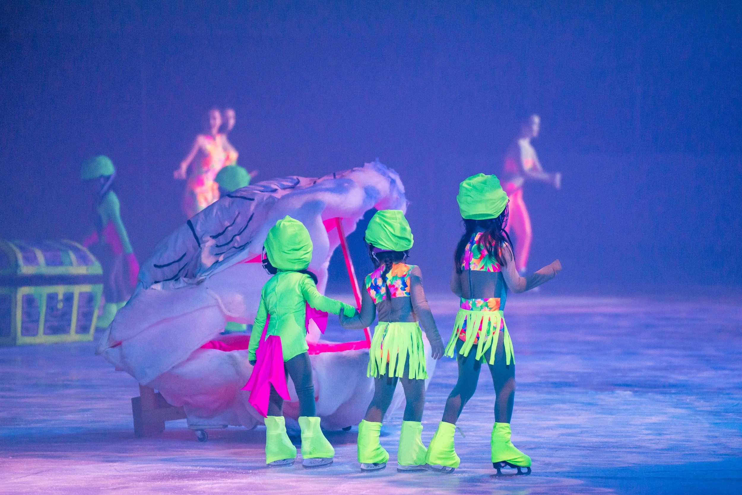 Ice skaters performing with a pink and white dragon prop on the ice, illuminated by vibrant, neon-colored lighting.