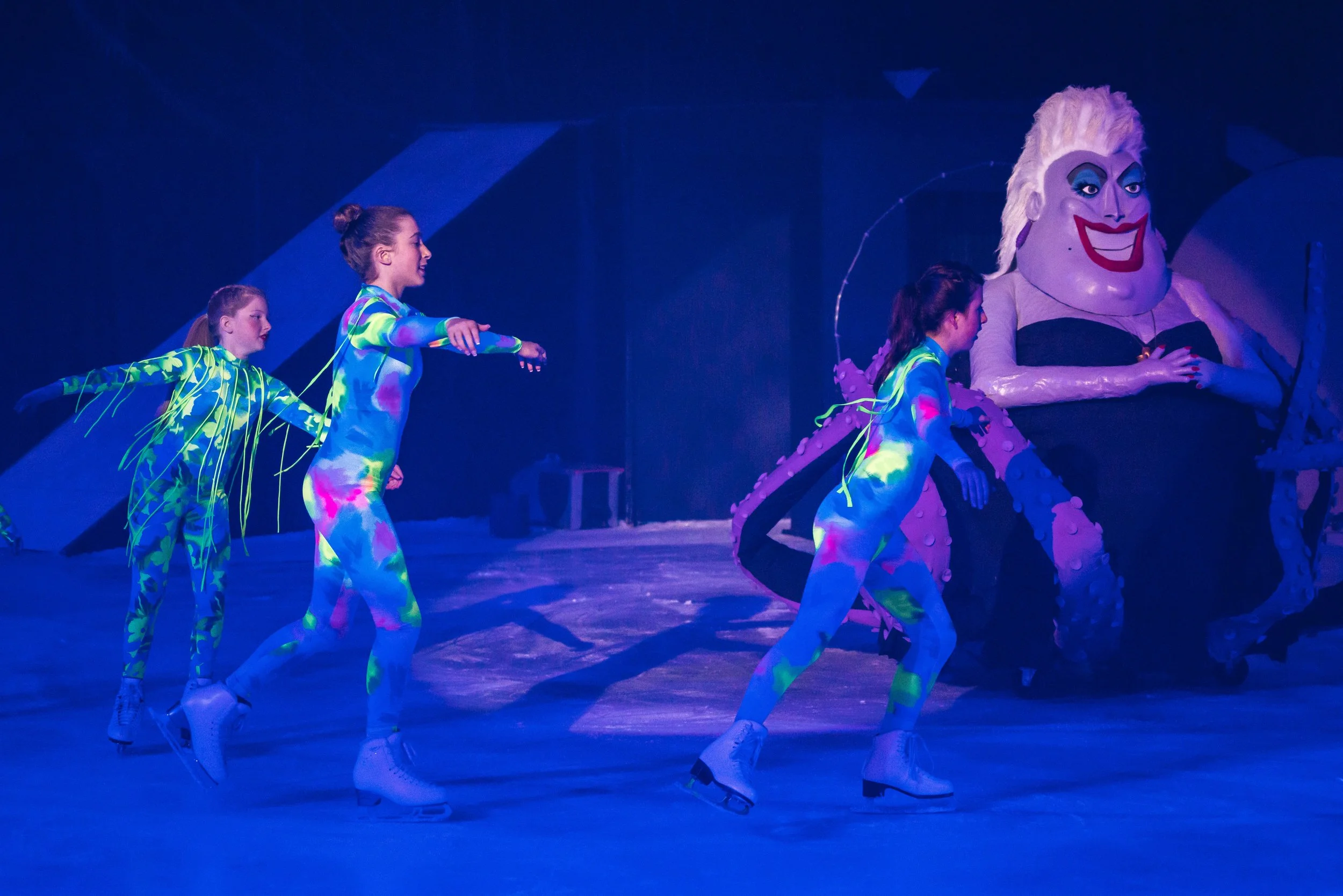Three young women in colorful, neon-patterned skating outfits perform synchronized ice skating routines in a rink under blue lighting, with a large clown figure in the background.