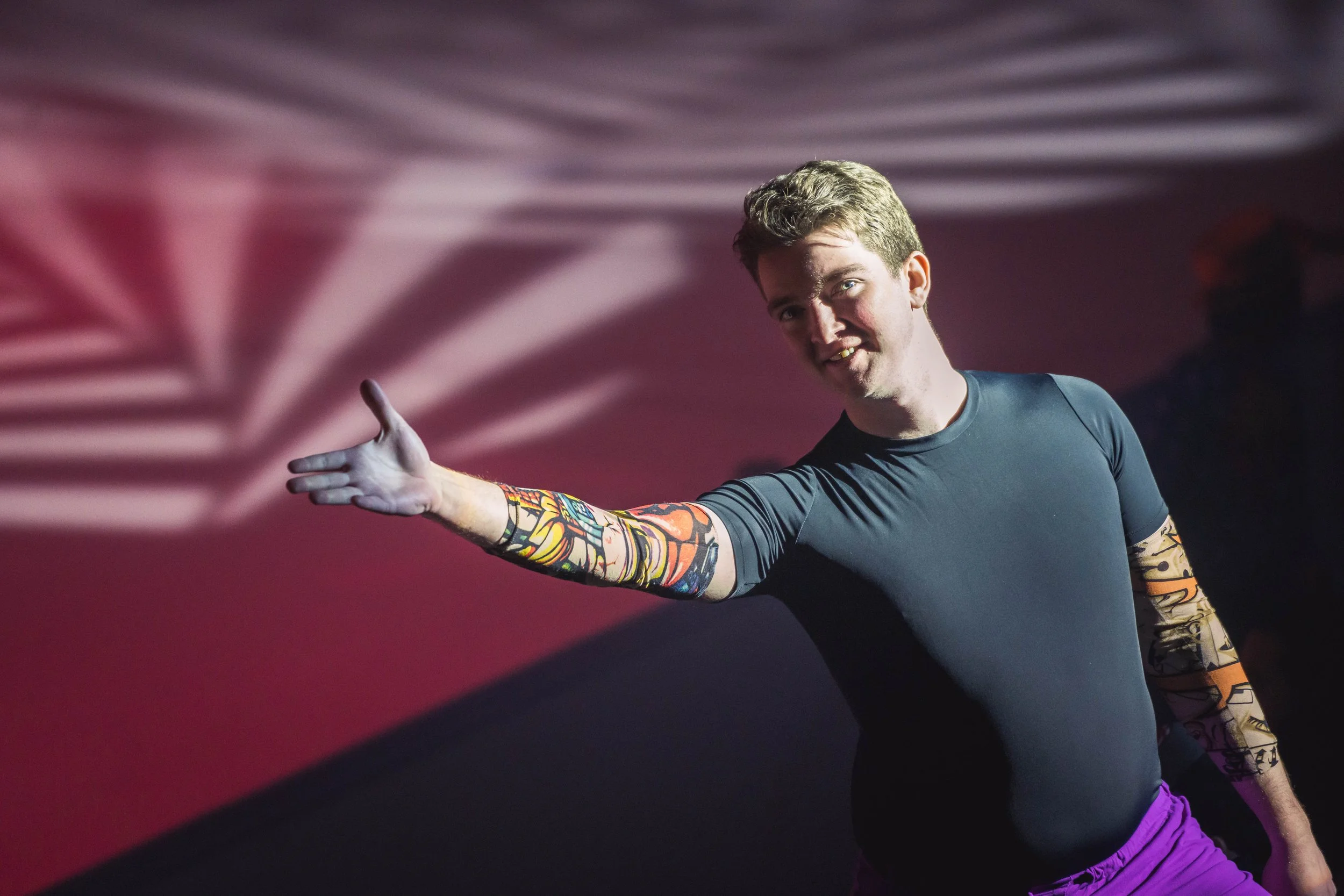 A young man with tattoos on his arm, wearing a dark athletic shirt and purple shorts, extends his arm outward with a friendly expression, standing in front of a background with wavy, pink and purple lines.