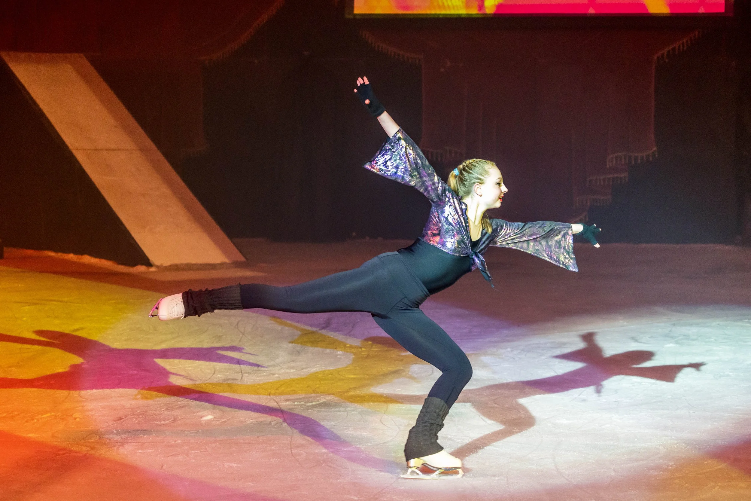 A female figure skater is performing a move on the ice rink, balancing on one leg with arms extended, dressed in a shiny, colorful costume and black ice skates.