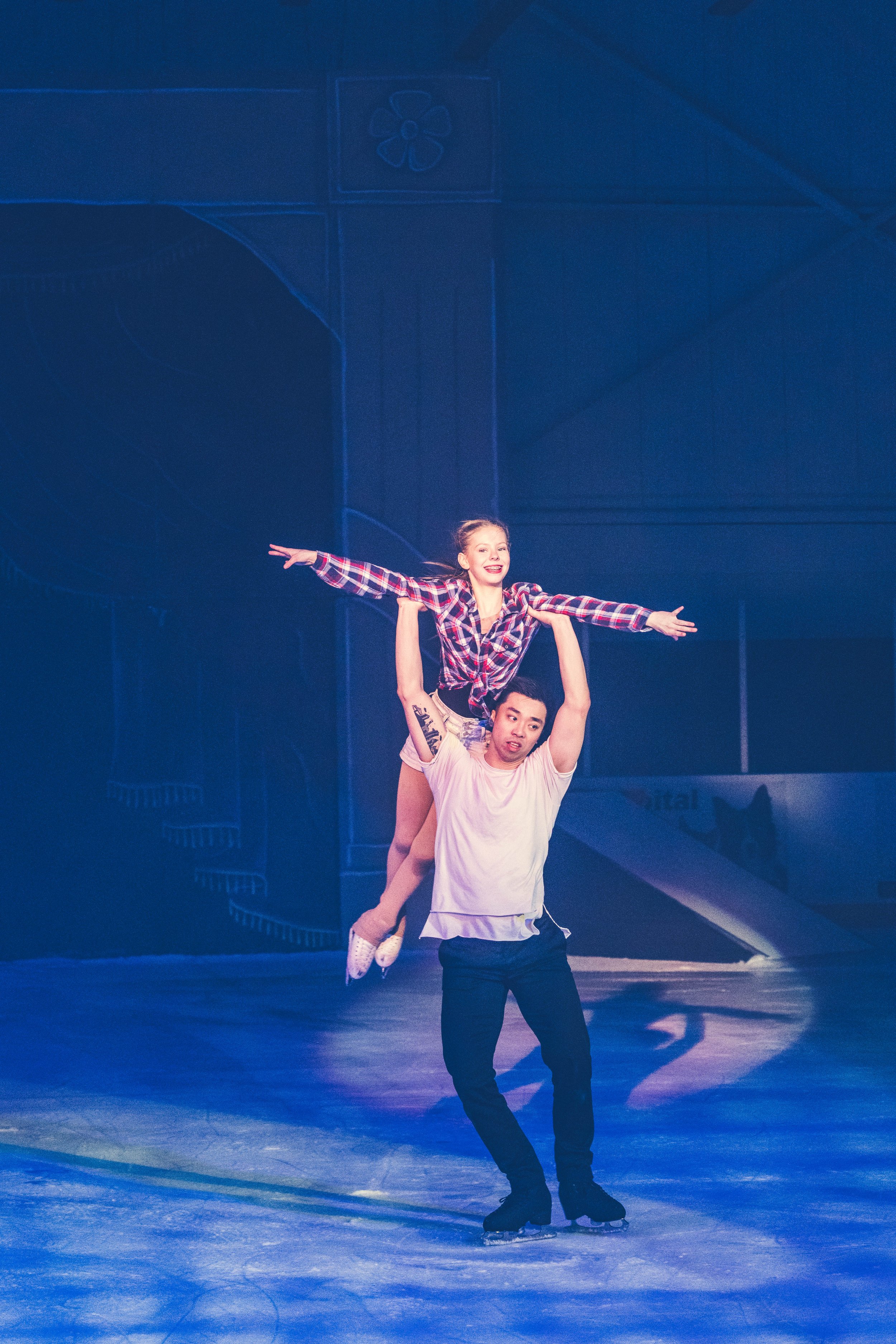 A male figure skater dressed in white gliding across the ice with a female partner sitting on his shoulders, arms outstretched, wearing a plaid shirt and shorts, in an ice skating rink.