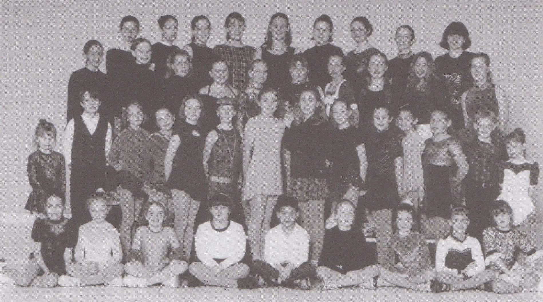 Black and white photo of a large group of young girls and women, arranged in five rows, posing for a group picture against a plain wall.