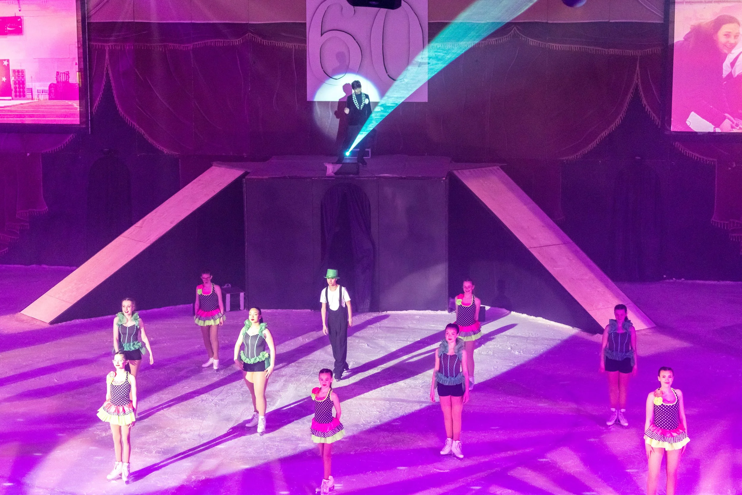 A stage with a group of children dressed in colorful costumes, performing a dance routine. The stage has purple lighting, and a performer dressed in black with a checkered sash is standing in front of a large whale illustration backdrop. Large screen
