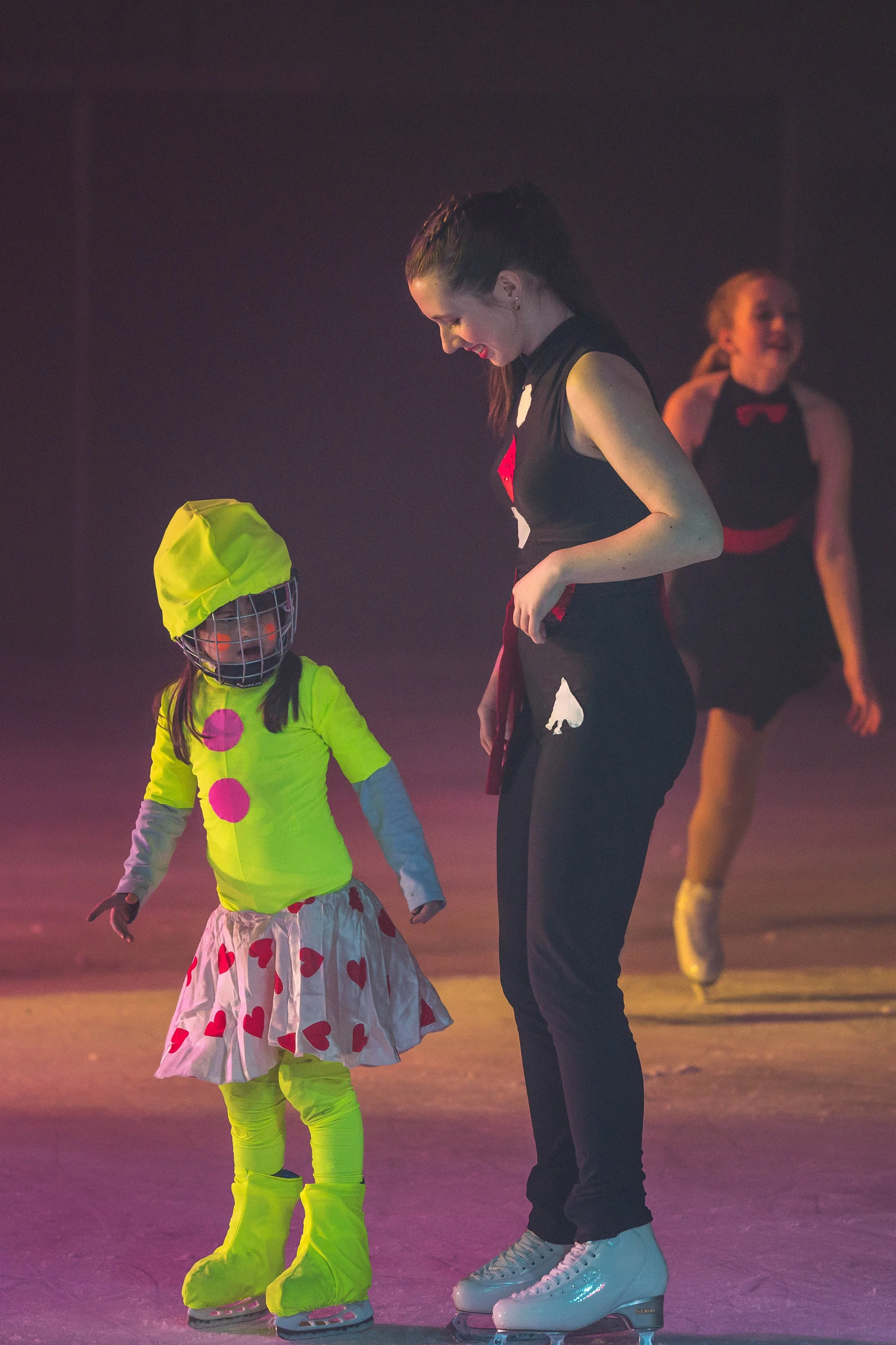 A young girl and an older woman ice skating together in a dimly lit arena. The girl is wearing a yellow hoodie with pink dots over a white dress with red hearts, and bright yellow ice skates. The woman is dressed in black, with her hair in a ponytail