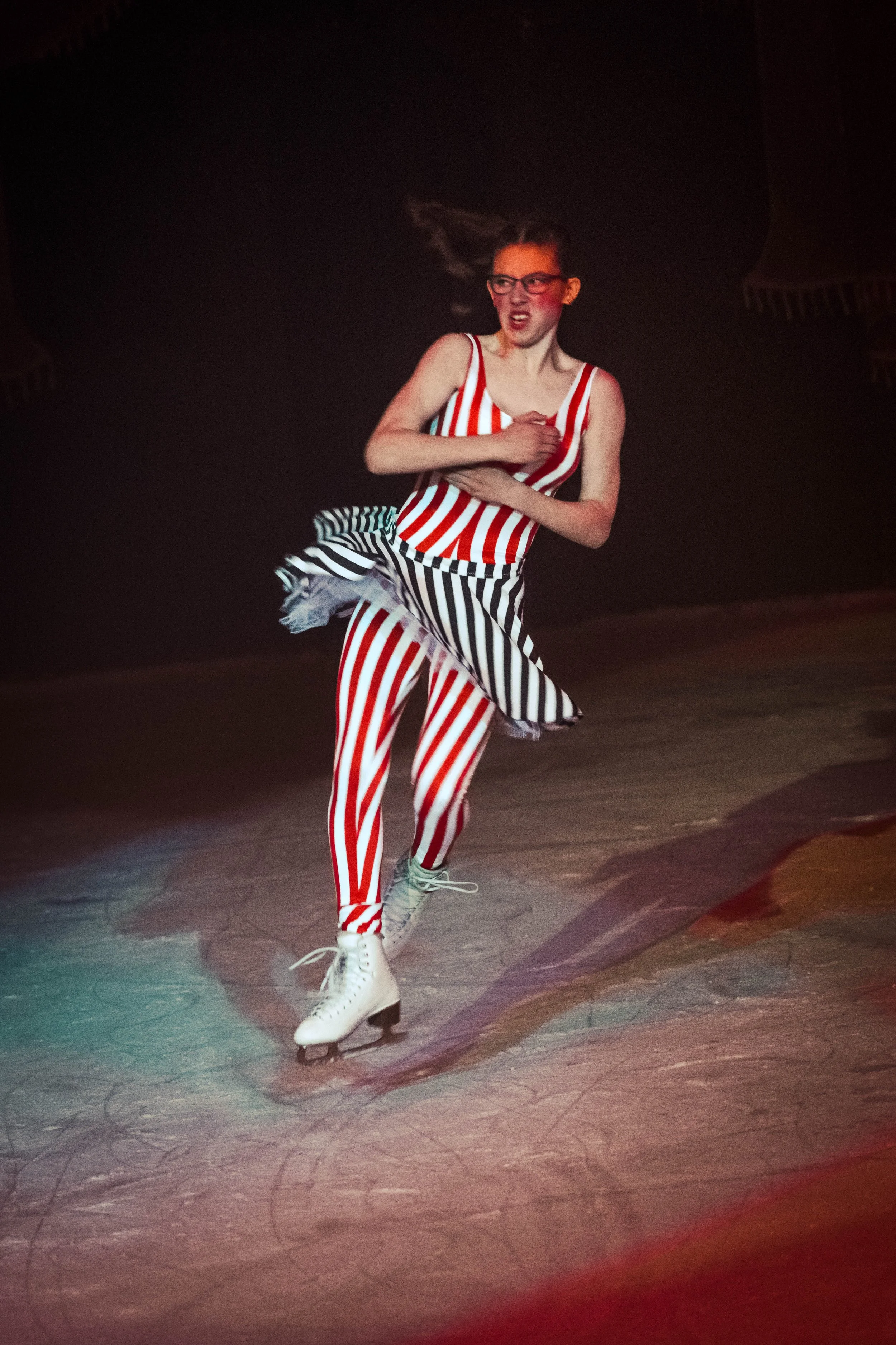 A young woman ice skating on an ice rink at night, wearing a red and white vertically striped tank top, matching red and white striped pants, and white ice skates. She has her hair in a ponytail and is making an expressive face, holding her chest wit