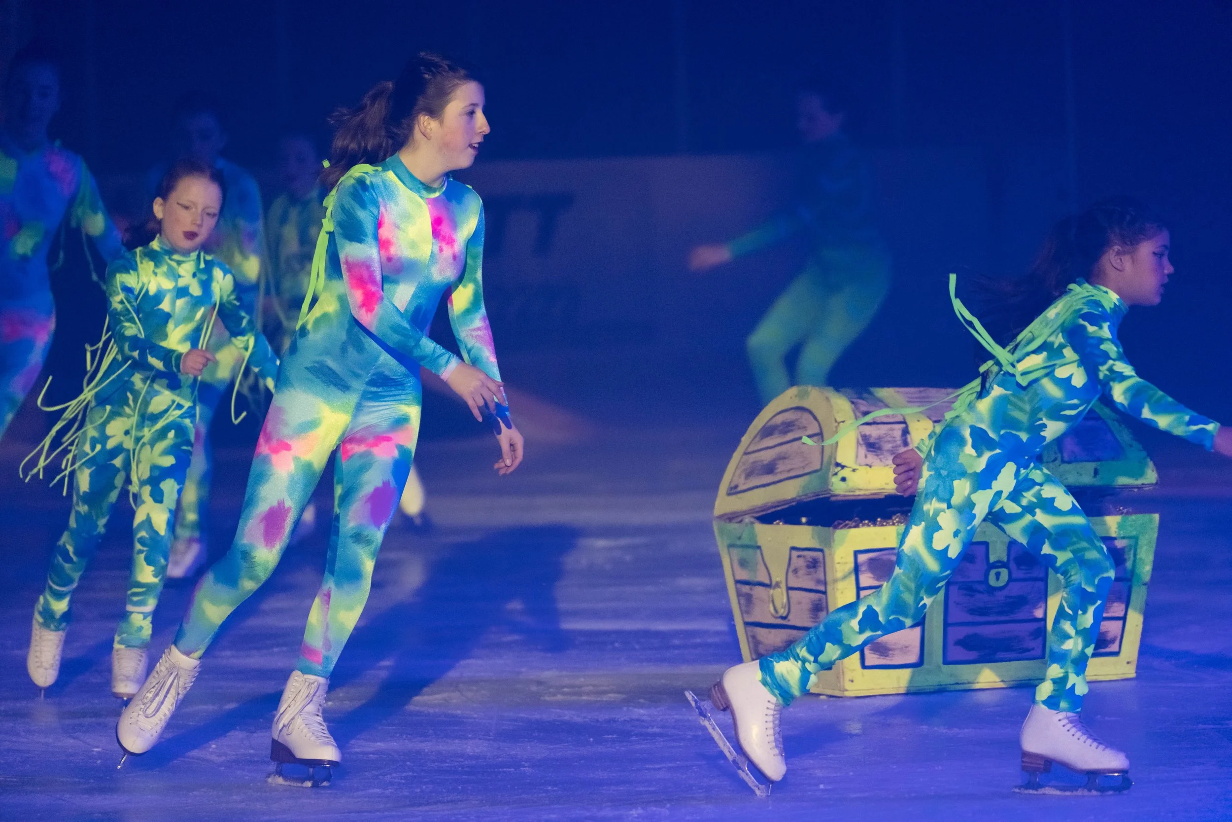 Ice skaters performing on stage wearing colorful, tie-dye costumes with yellow laces. One girl is pulling a painted cardboard treasure chest prop, and others are skating behind her.