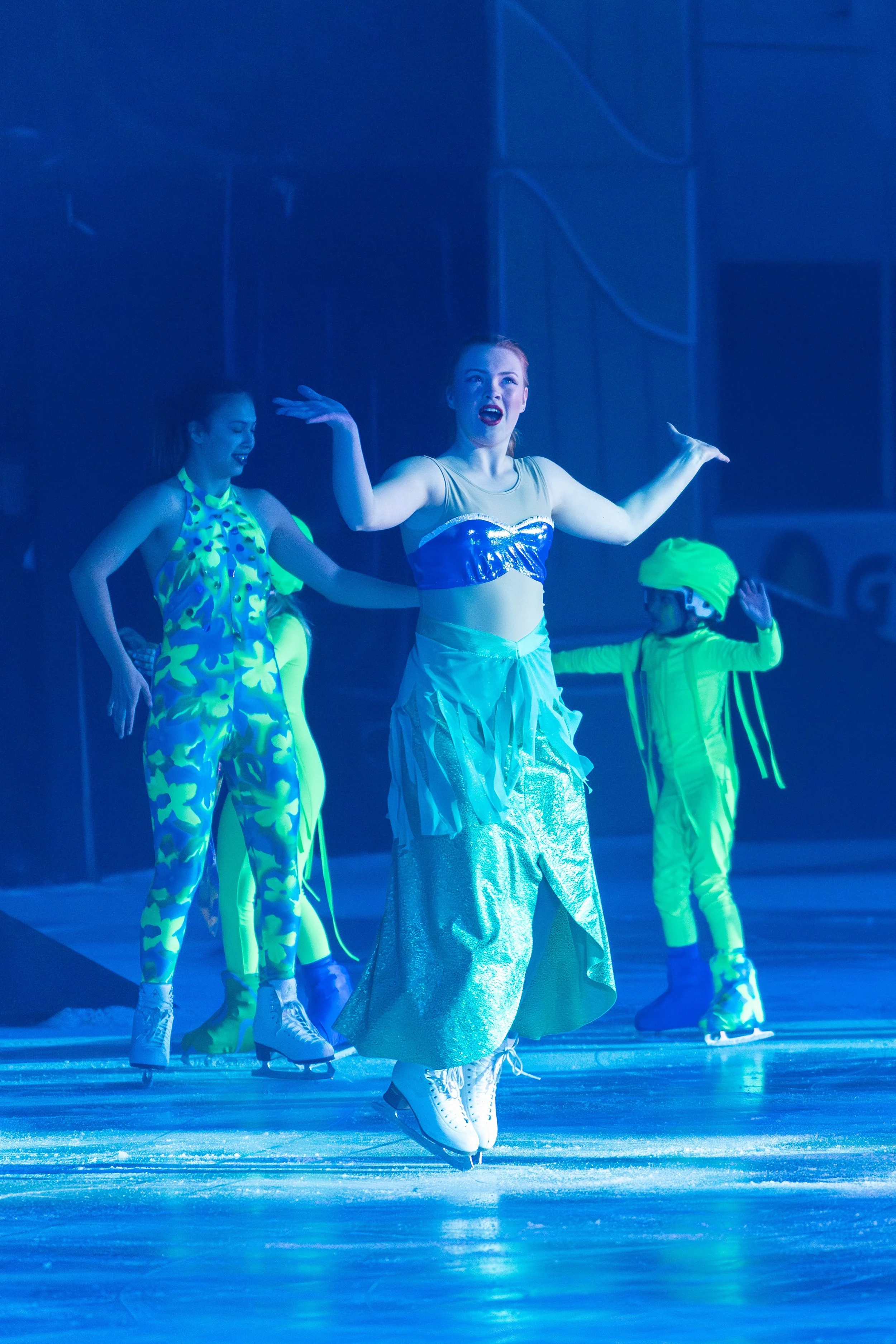Skaters performing on ice, illuminated in blue lighting, with a female skater in metallic attire and colorful costumes of others visible in the background.
