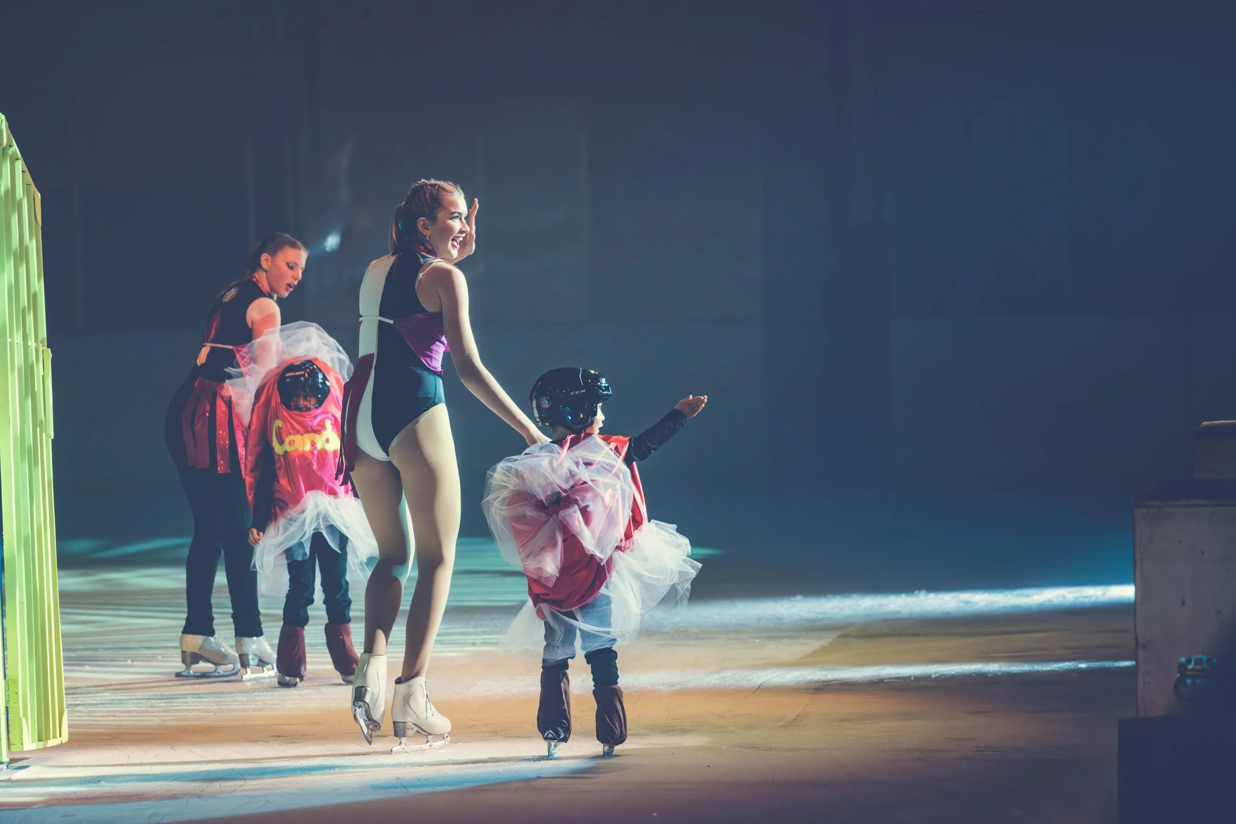 A young woman figure skater wearing a black, pink, and purple costume and white ice skates skating on ice, smiling and waving at children dressed as princesses with pink tutus and tiaras, holding hands in a line inside an ice rink.