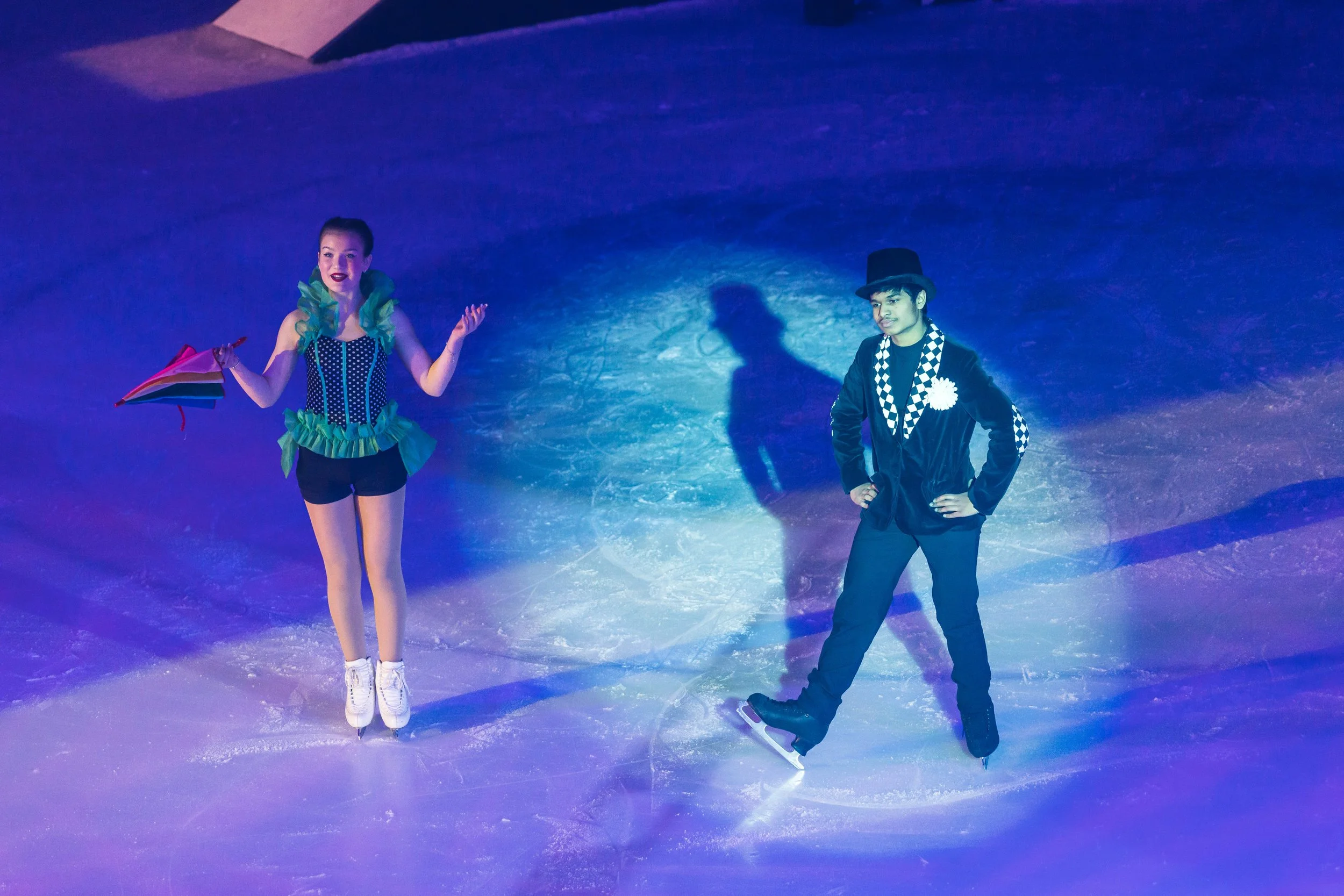 Two figure skaters standing on an ice rink under colorful lighting. The female skater is skating with her arms raised and holding a small rainbow-colored flag, wearing a black and green costume. The male skater stands with hands on hips, wearing a to