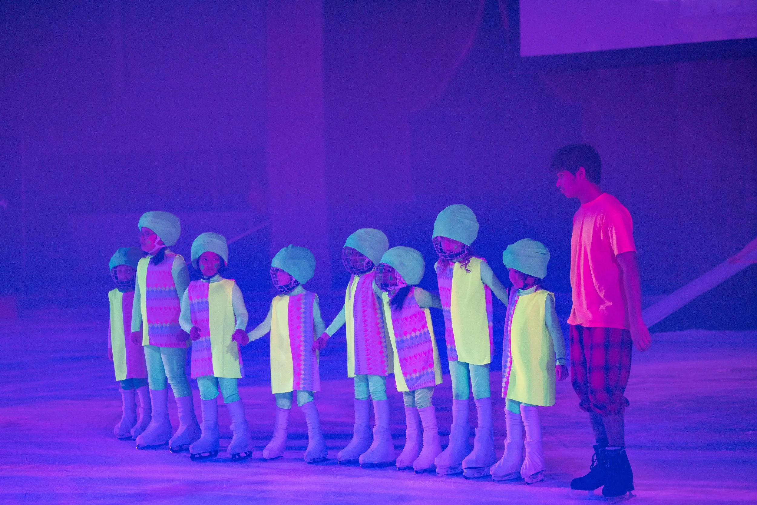 A group of children wearing white hats and outfits lined up holding hands, with a person in a pink shirt standing next to them on a stage illuminated with purple light.