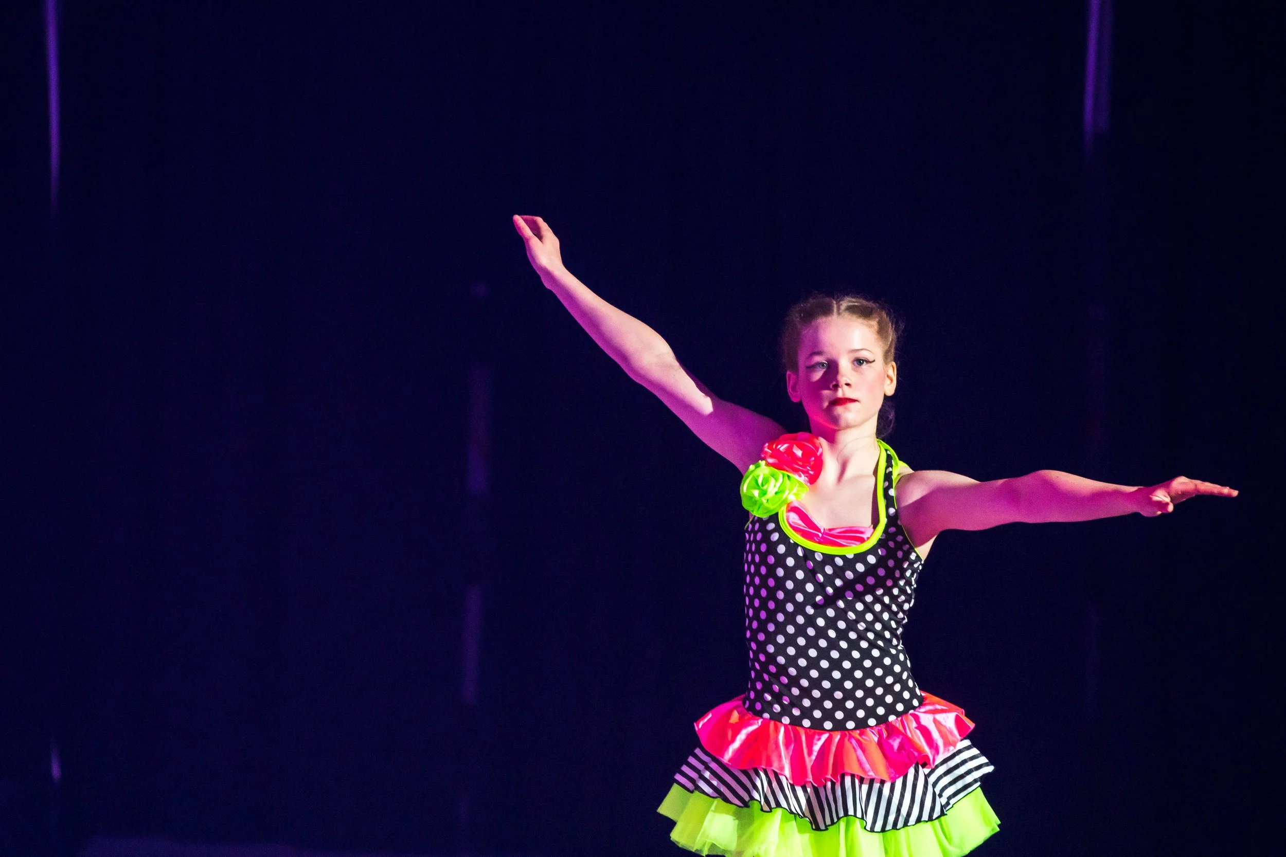 Young girl in a colorful polka dot and striped costume performing a dance on stage with dark background.
