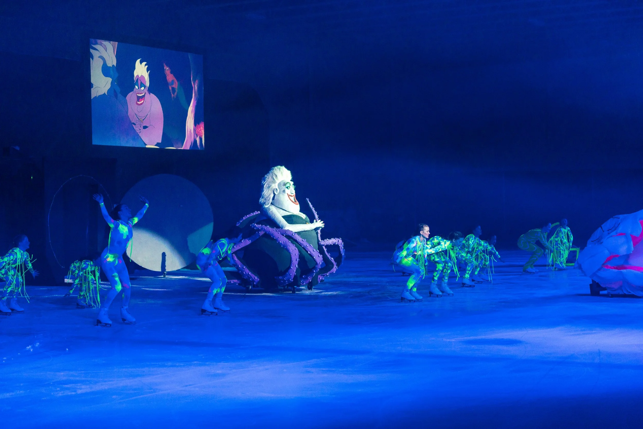 A vibrant stage performance featuring Disney's Ursula from The Little Mermaid, surrounded by dancers in luminous costumes under blue lighting, with a large screen displaying animated scenes of Ursula in the background.