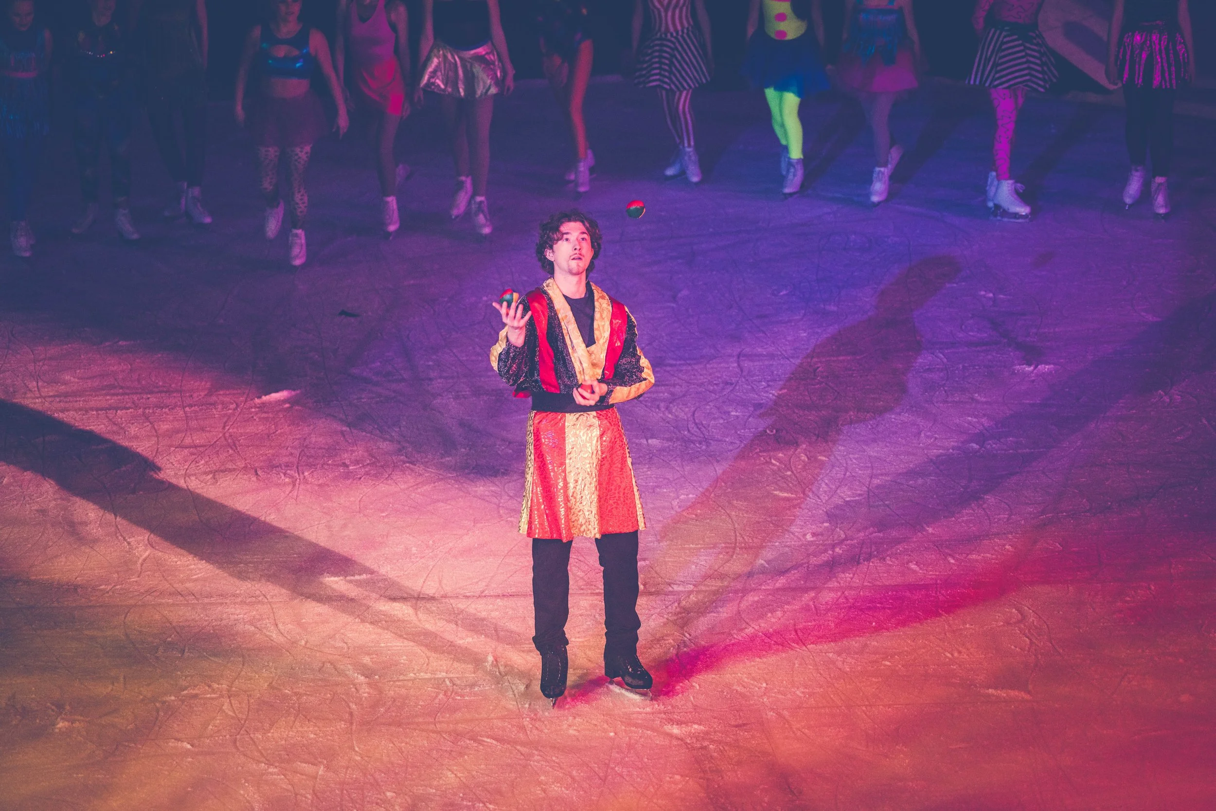 A man wearing a multicolored robe figure skates and juggles on an ice rink, while a group of women in colorful costumes and ice skates stand behind him.