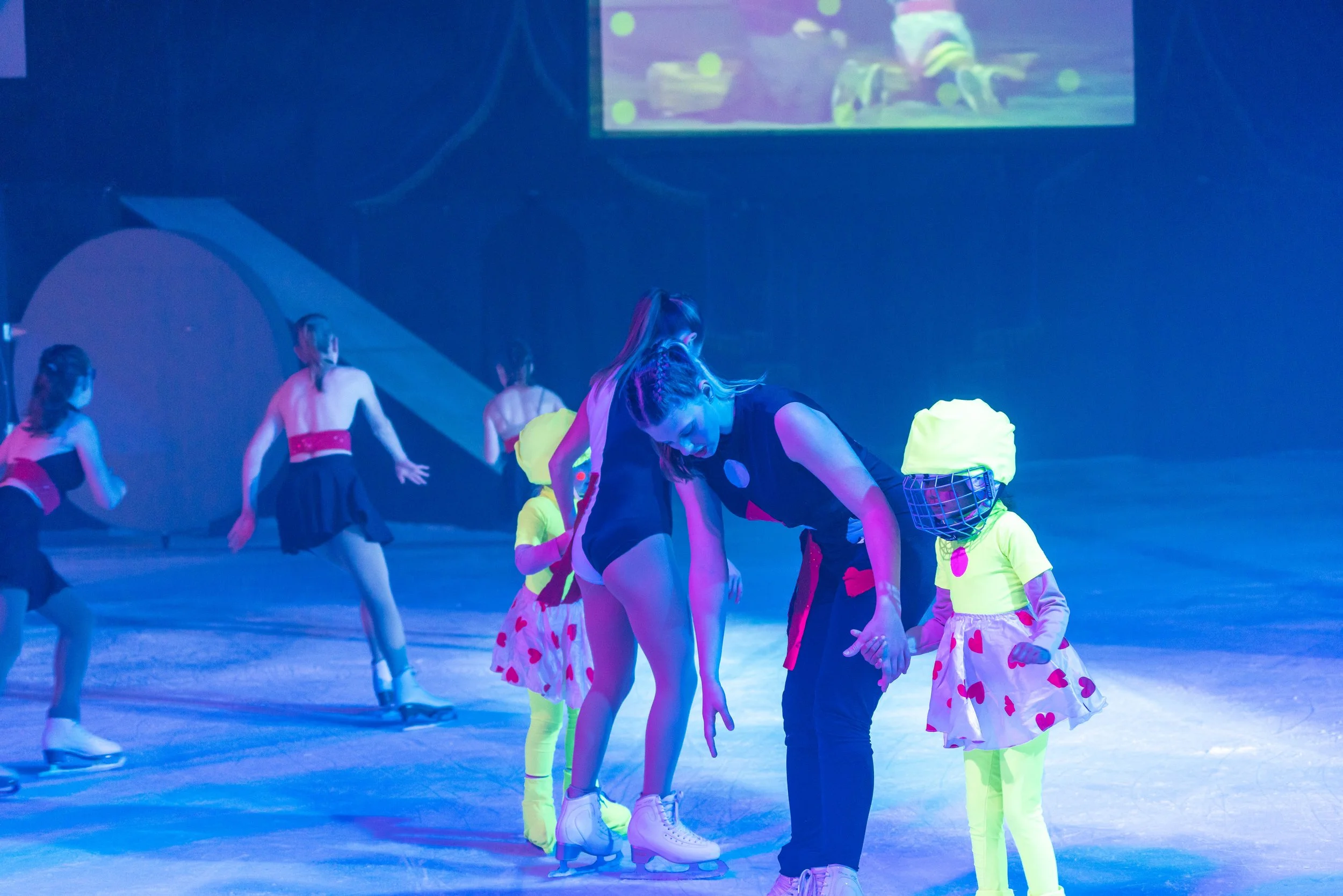 A group of children and a young woman dressed in colorful costumes with heart patterns, skating on an ice rink under blue lighting. Some children are wearing bright yellow or neon green outfits with plaid or heart patterns, and the young woman in bla