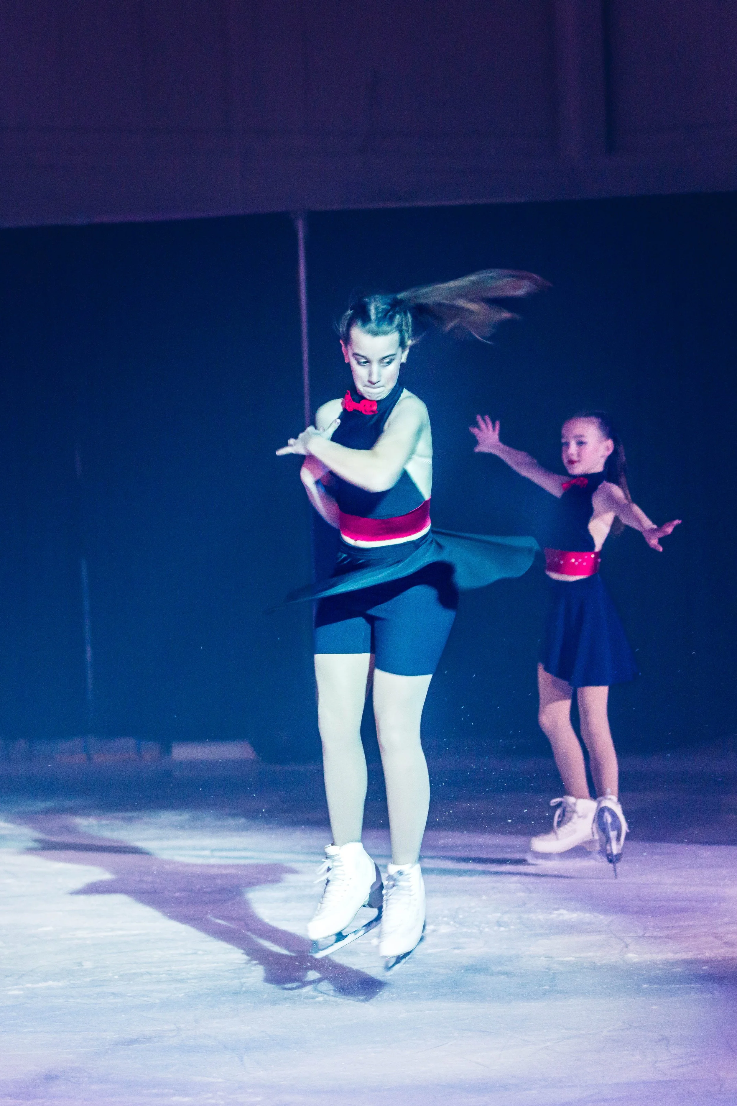 Two young female ice skaters performing on an ice rink, wearing black costumes with red accents, one in a skirt and the other in leggings, with the main skater in the foreground executing a leap or spin.