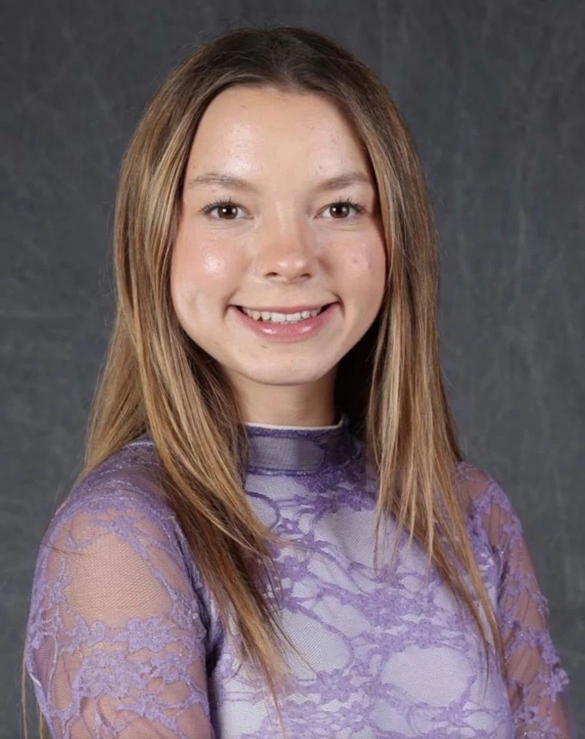 A young woman with light skin, shoulder-length light brown hair, smiling, wearing a purple lace top, standing against a gray background.