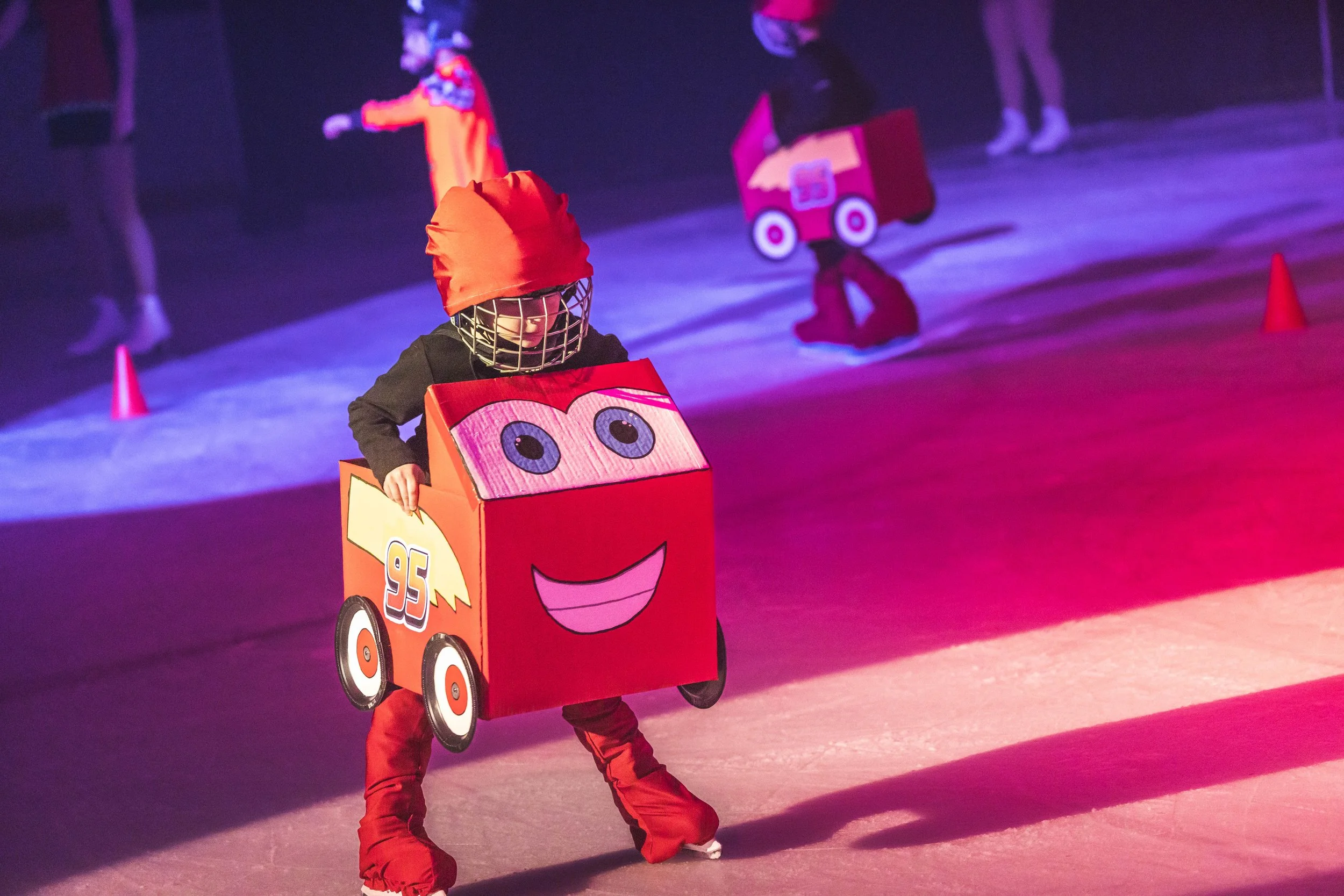 Child dressed in a Lightning McQueen car costume at an indoor skating rink, surrounded by cones and other children skating in the background.