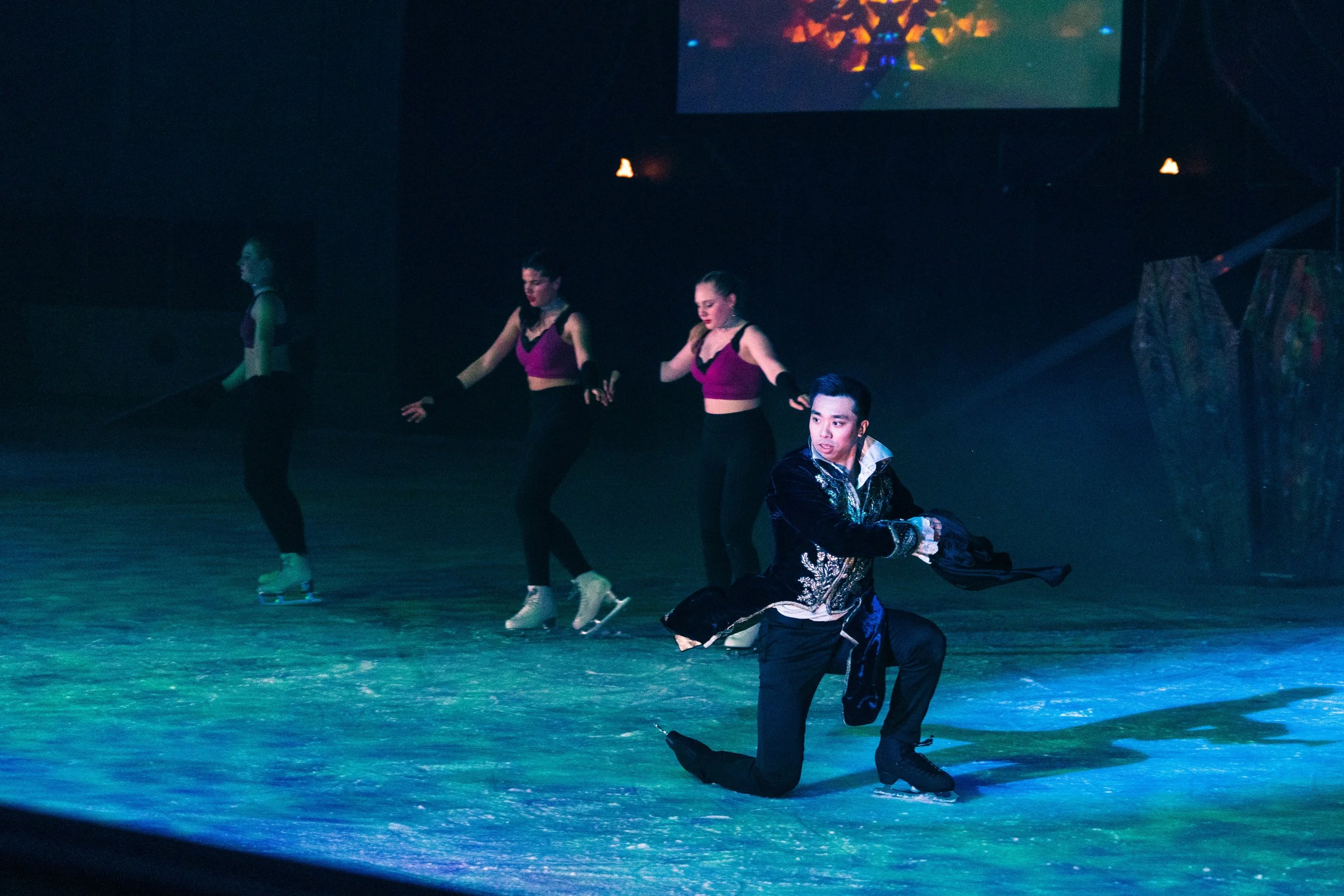 A group of figure skaters performing on an ice rink with colorful stage lighting and a digital screen in the background.