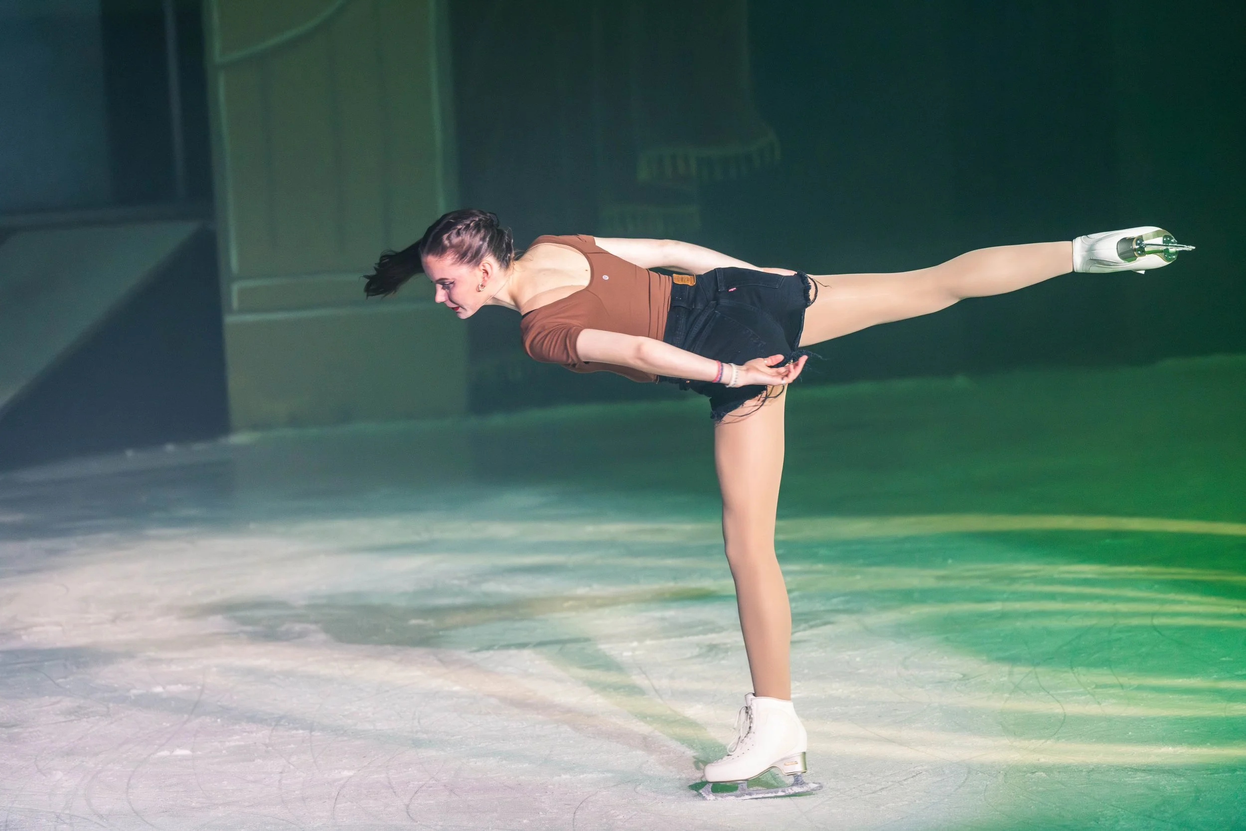 A female figure skater performing a move on the ice rink, wearing a brown top, black shorts, and white figure skates, with her hair styled in a braid.