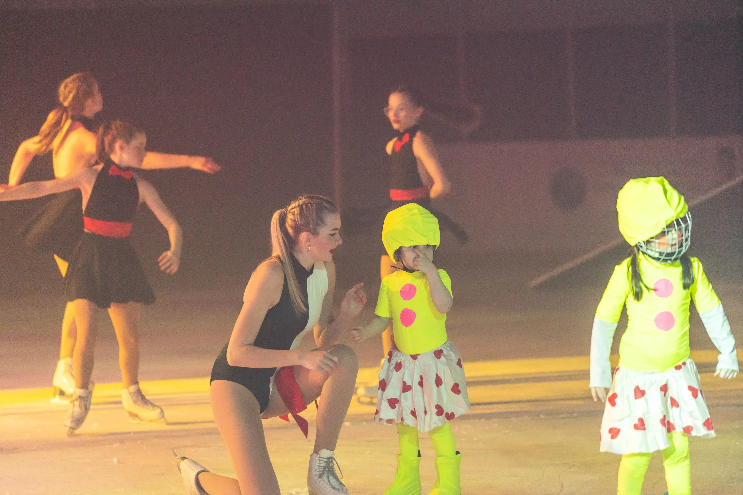 Performers in a dance or theatrical production, including children dressed as clowns and young women in black costumes, on stage with a foggy background and stage lighting.