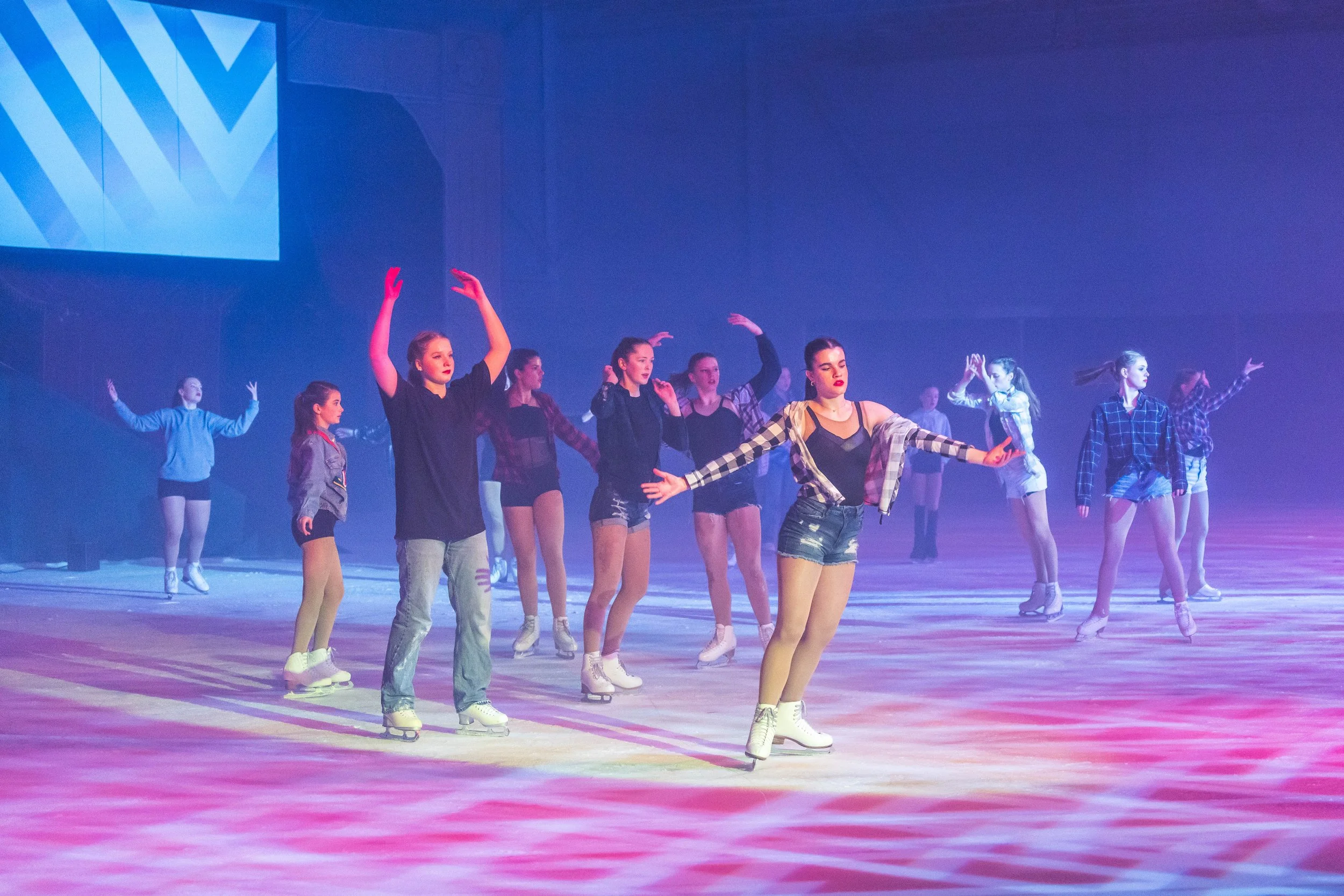 Group of young women and girls wearing ice skates practicing ballet or dance routines on an ice rink under colorful stage lighting.