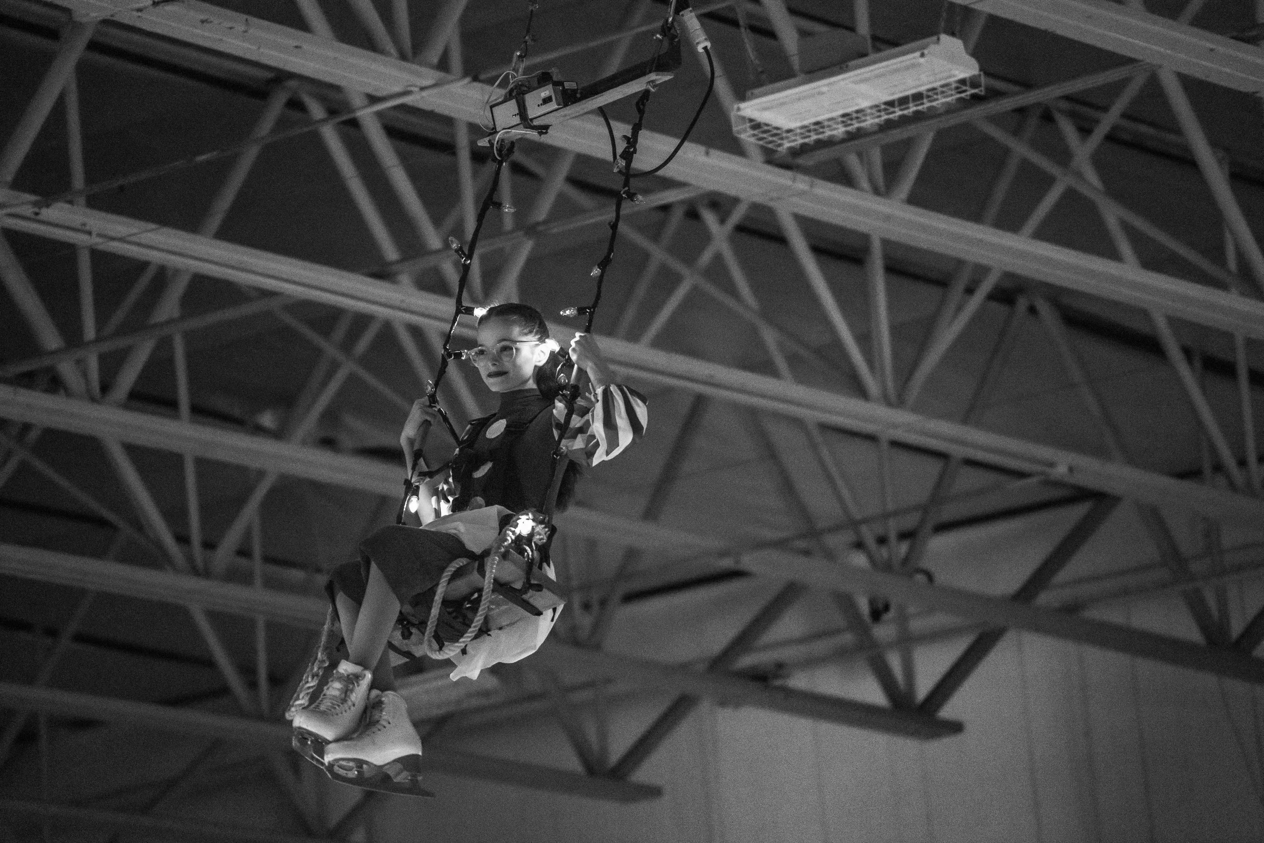 A young girl with glasses, wearing a polka dot top, is riding a swing suspended from a metal framework at an indoor amusement park or play area.