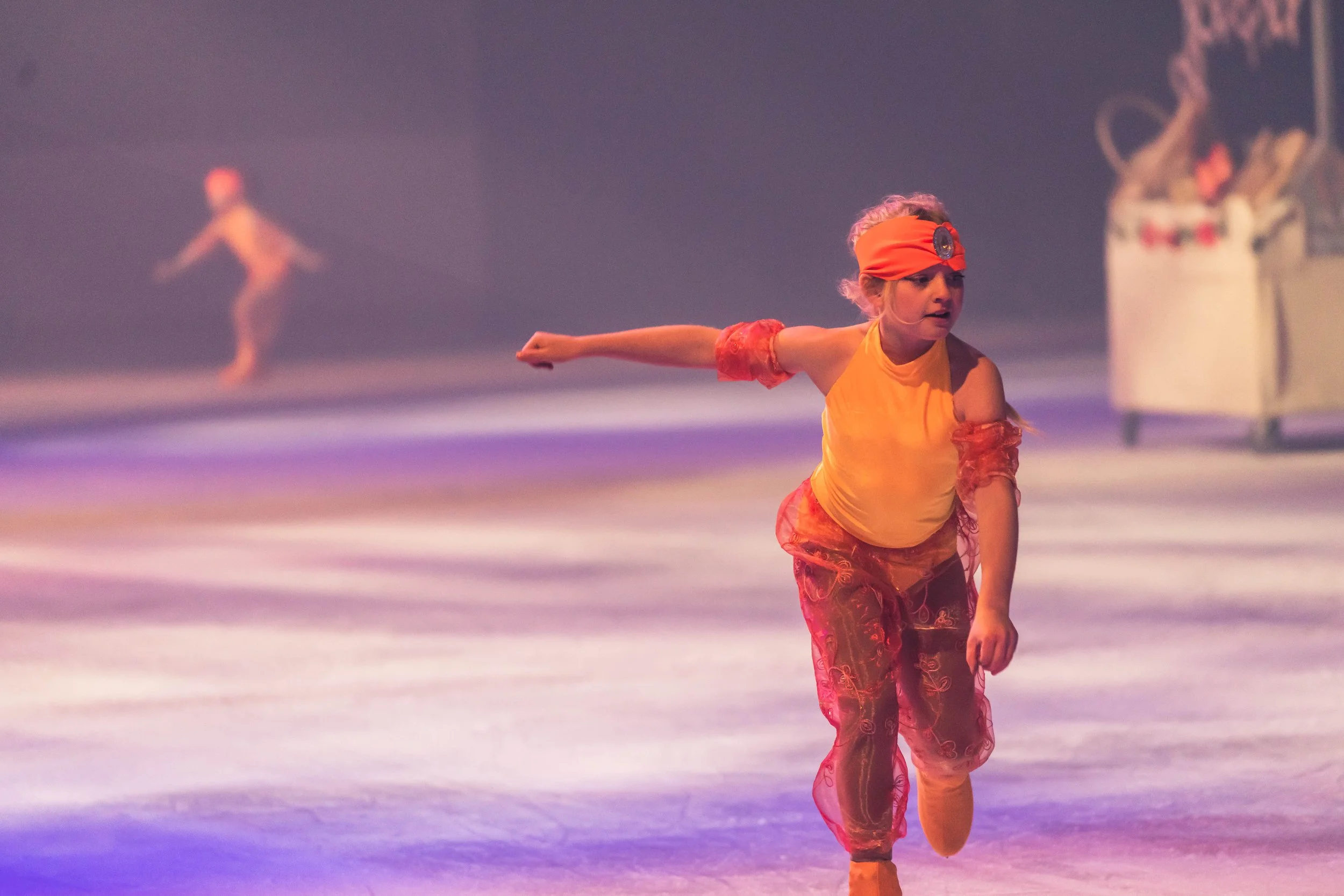 A young girl dressed in orange performing a dance move on ice, with a blurred figure in similar attire in the background.
