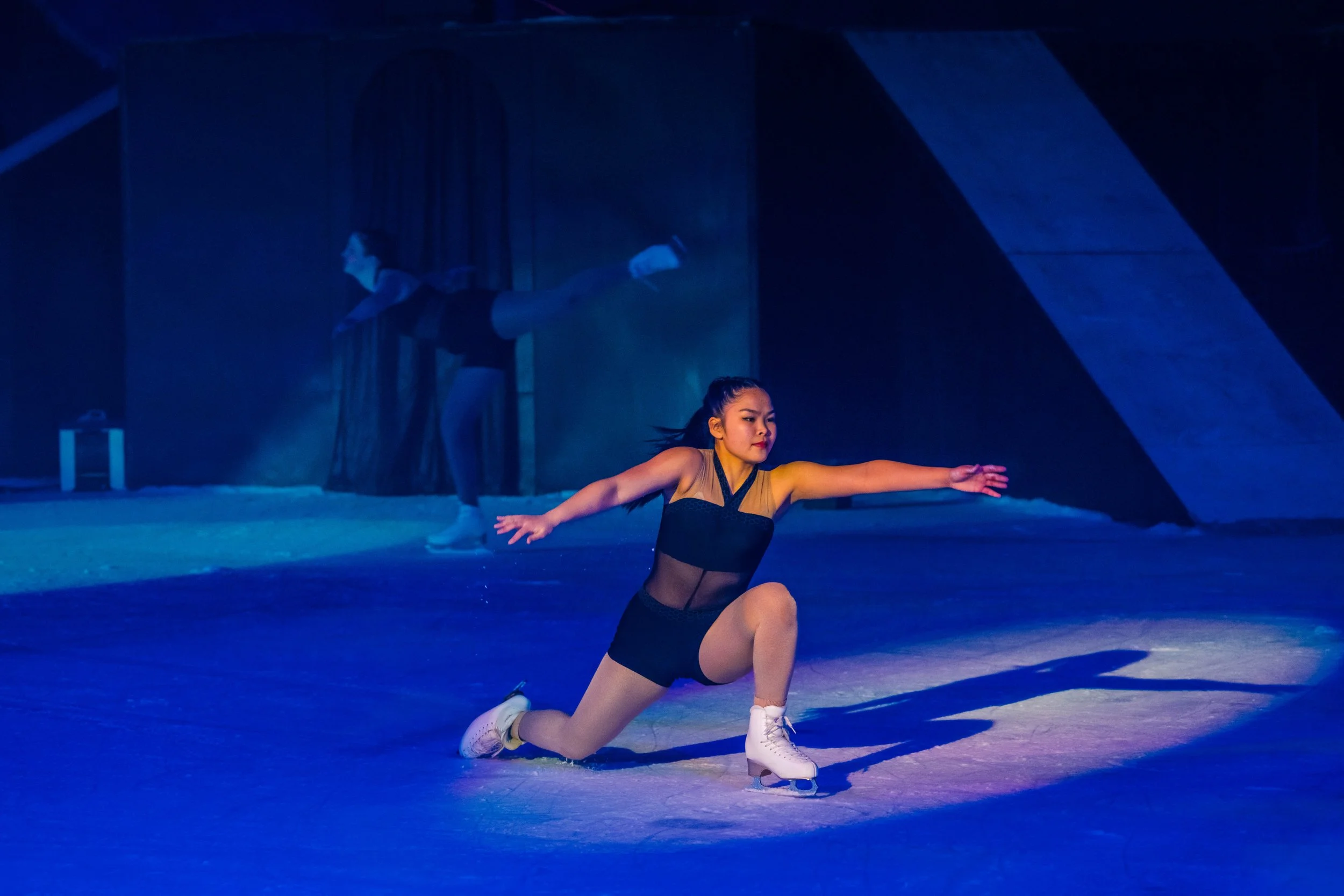 A female figure skater performing a move on the ice rink with her left knee on the ground and her arms extended, wearing a black and gold costume with ice skates, in a dimly lit setting with blue and purple lighting.