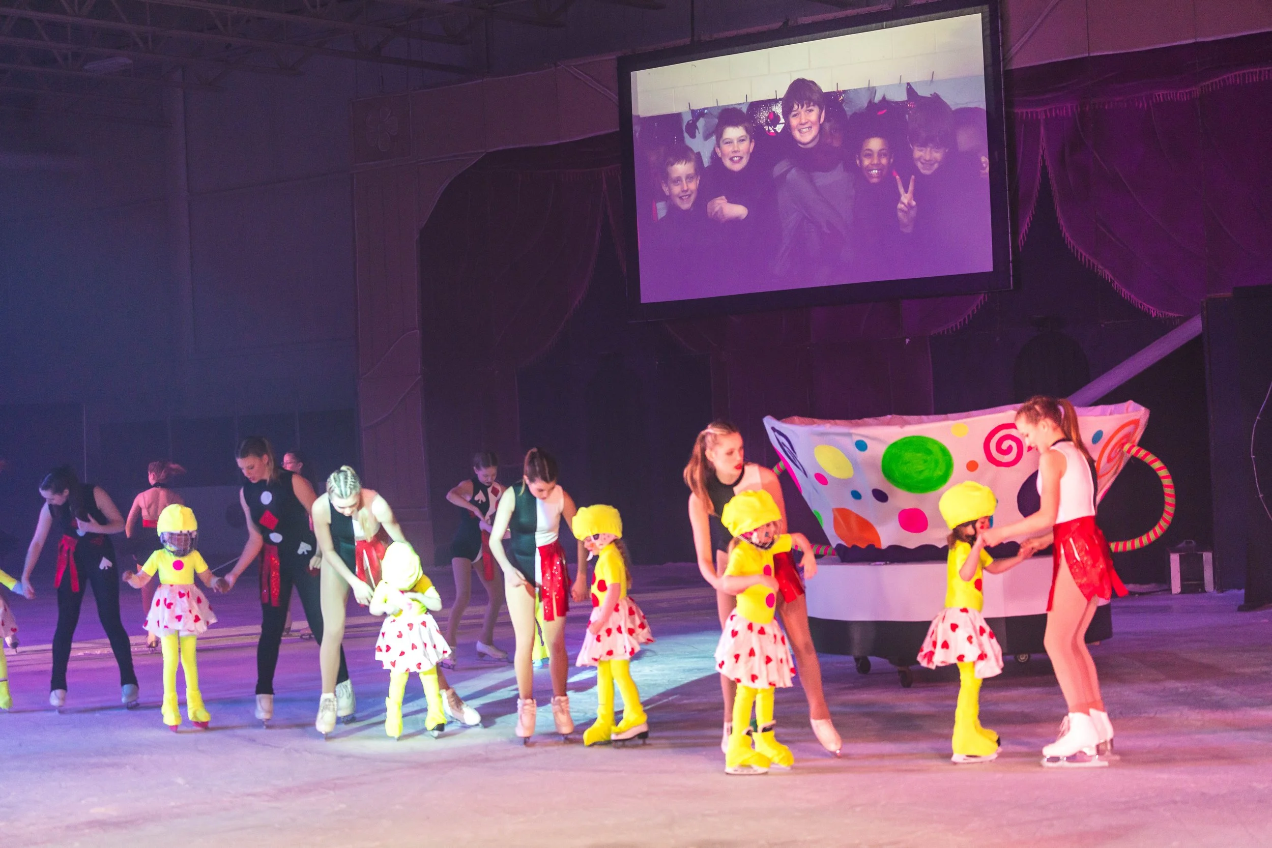 Children and teenagers performing a dance or skit on stage with a colorful polka dot backdrop, under purple stage lighting, with a large screen displaying a group photo of kids in the background.