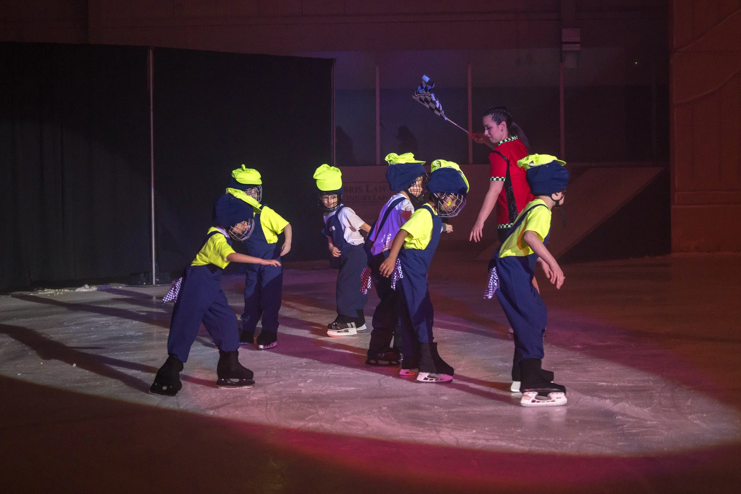 A group of young children wearing ice skates, yellow and blue uniforms with helmets, are gathered on an ice rink, họcing from a female instructor. The instructor is holding a hockey stick and wears a red shirt.